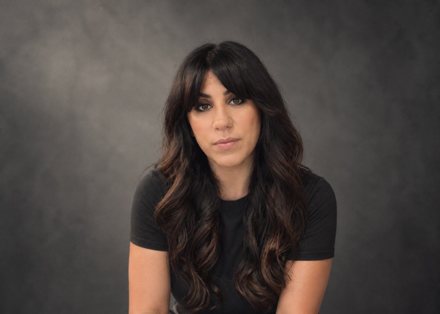 Portrait of a woman with long dark wavy hair and side-swept bangs, wearing a black t-shirt, against a gray background.