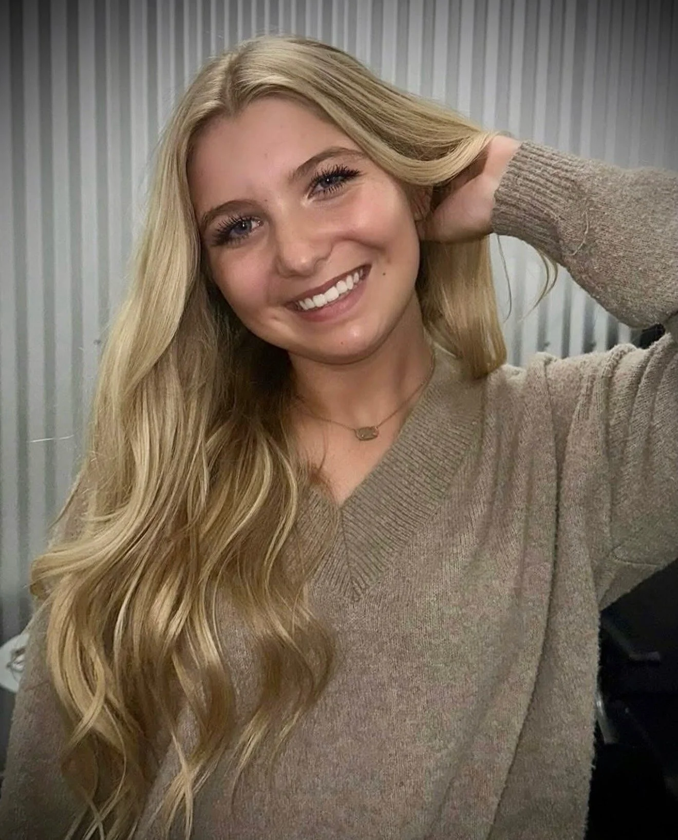 A young woman with long blonde hair smiling, wearing a beige sweater and a necklace, touching her hair with her right hand, in front of a gray metal wall.