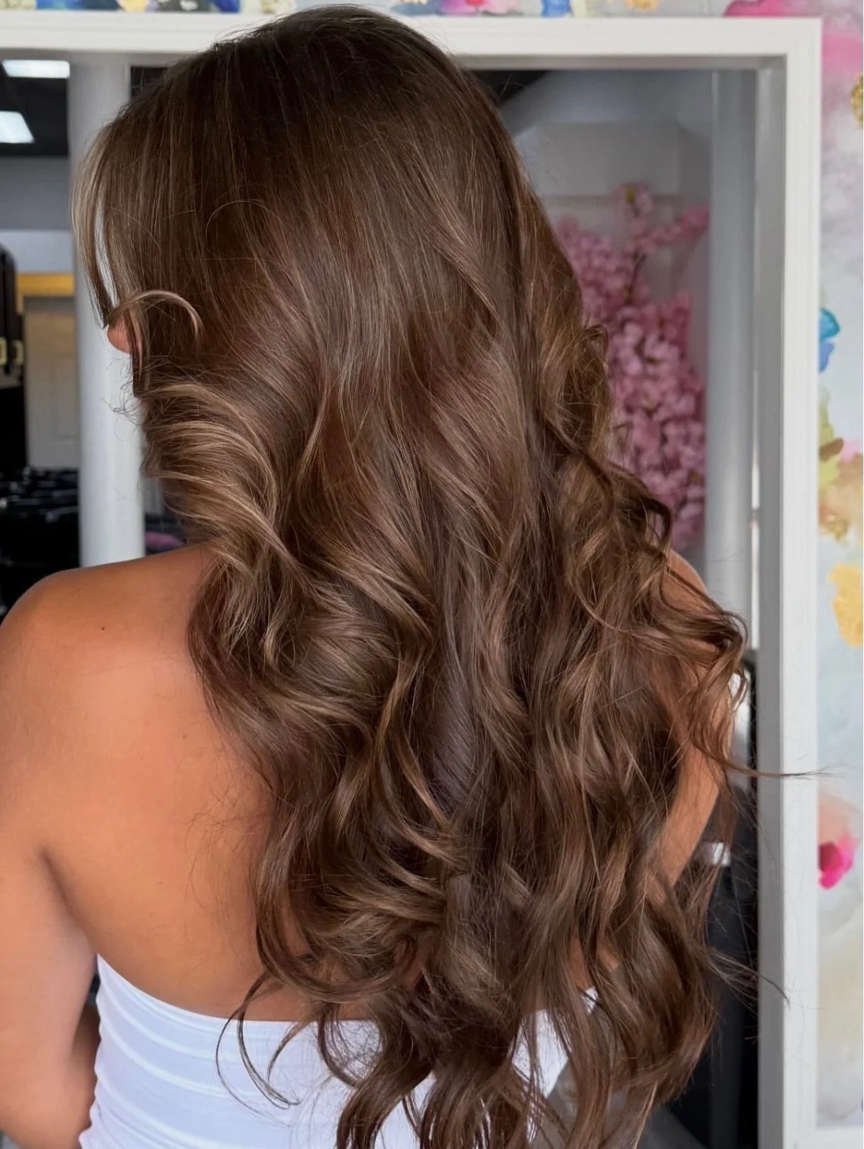 A woman with long, curly brown hair wearing a white tank top, seen from the back, standing indoors with a floral background.