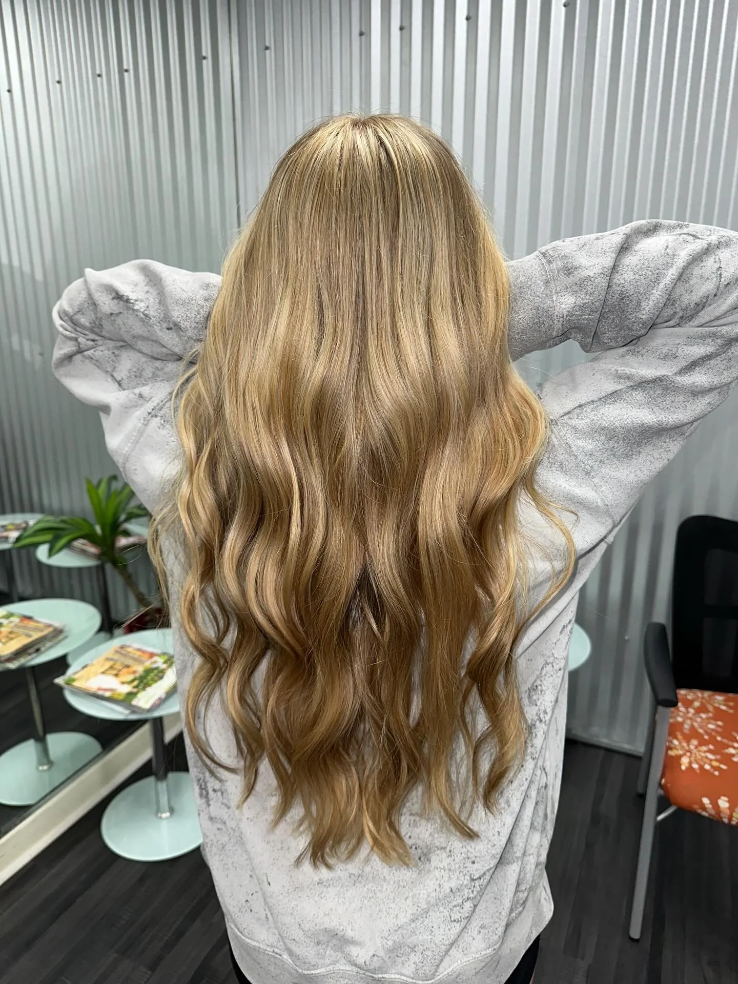 A woman with long wavy blonde hair seen from behind, with arms raised behind her head, wearing a light-colored sweatshirt in a room with gray walls, chairs, and tables.
