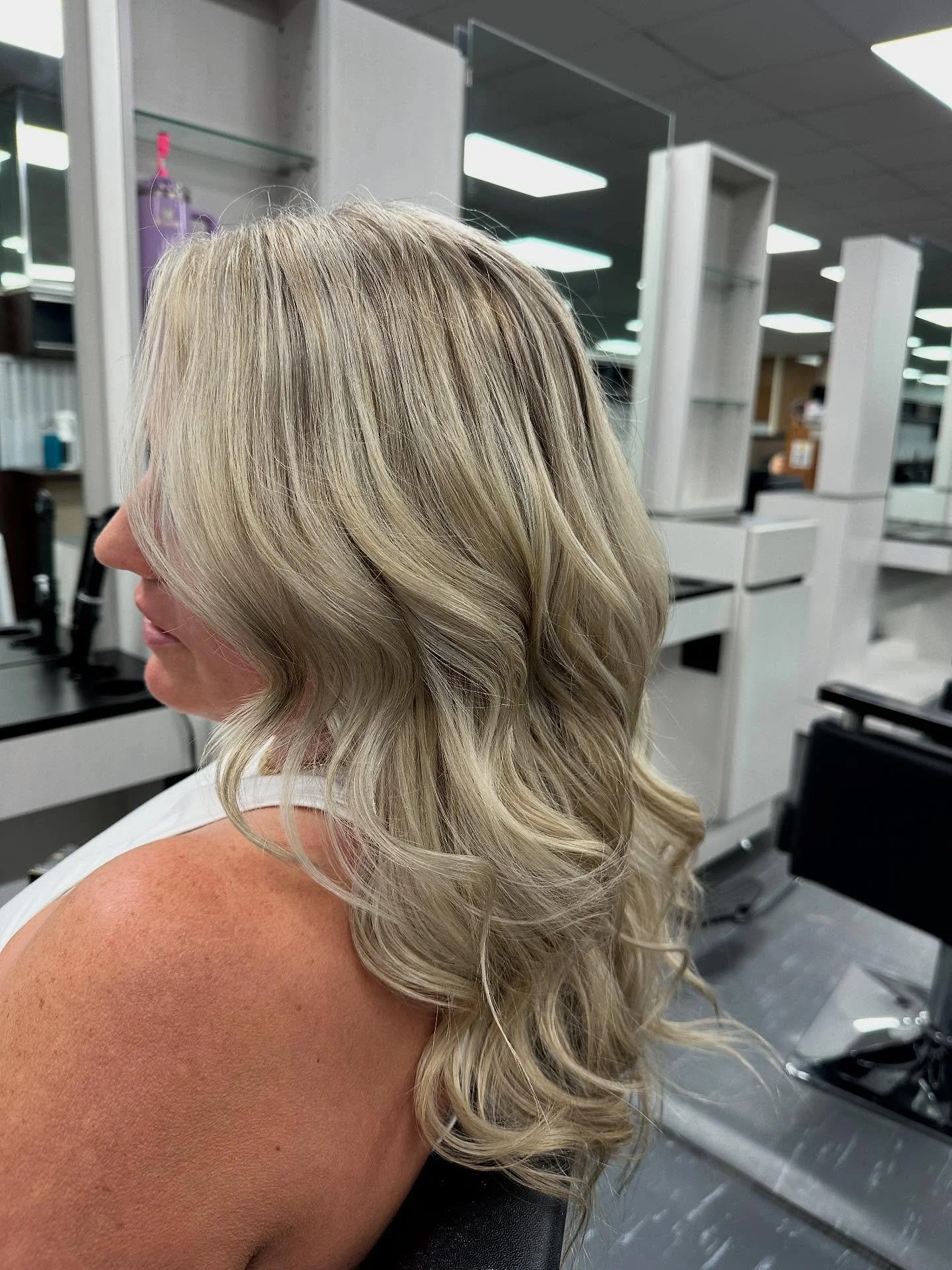 A woman with wavy blonde hair sitting in a salon, with hair styling tools and shelves in the background.