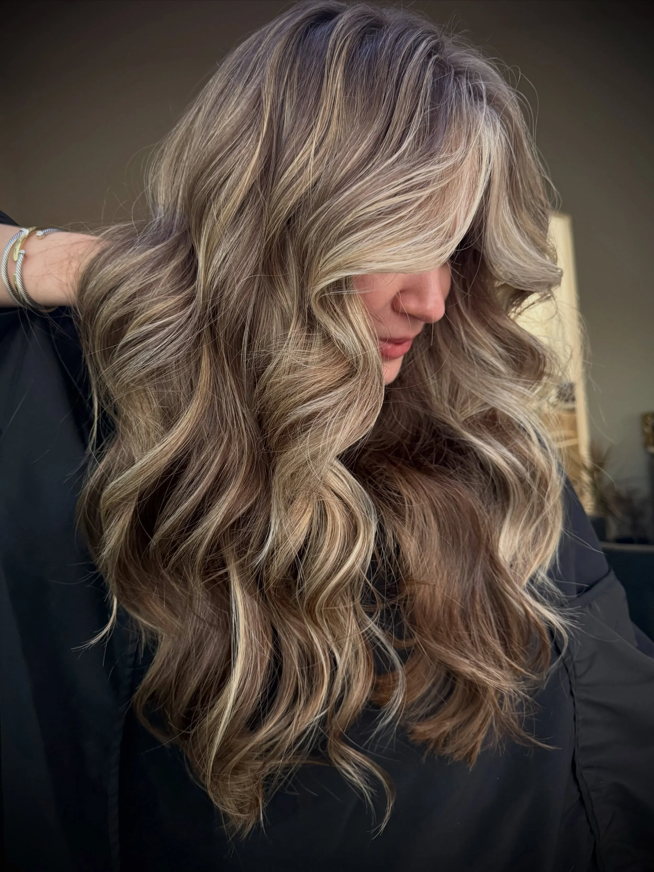 A woman with long, wavy, blonde and light brown hair, leaning her head down and resting her hand on her neck, wearing several rings and a black top. The background is dark and out of focus.
