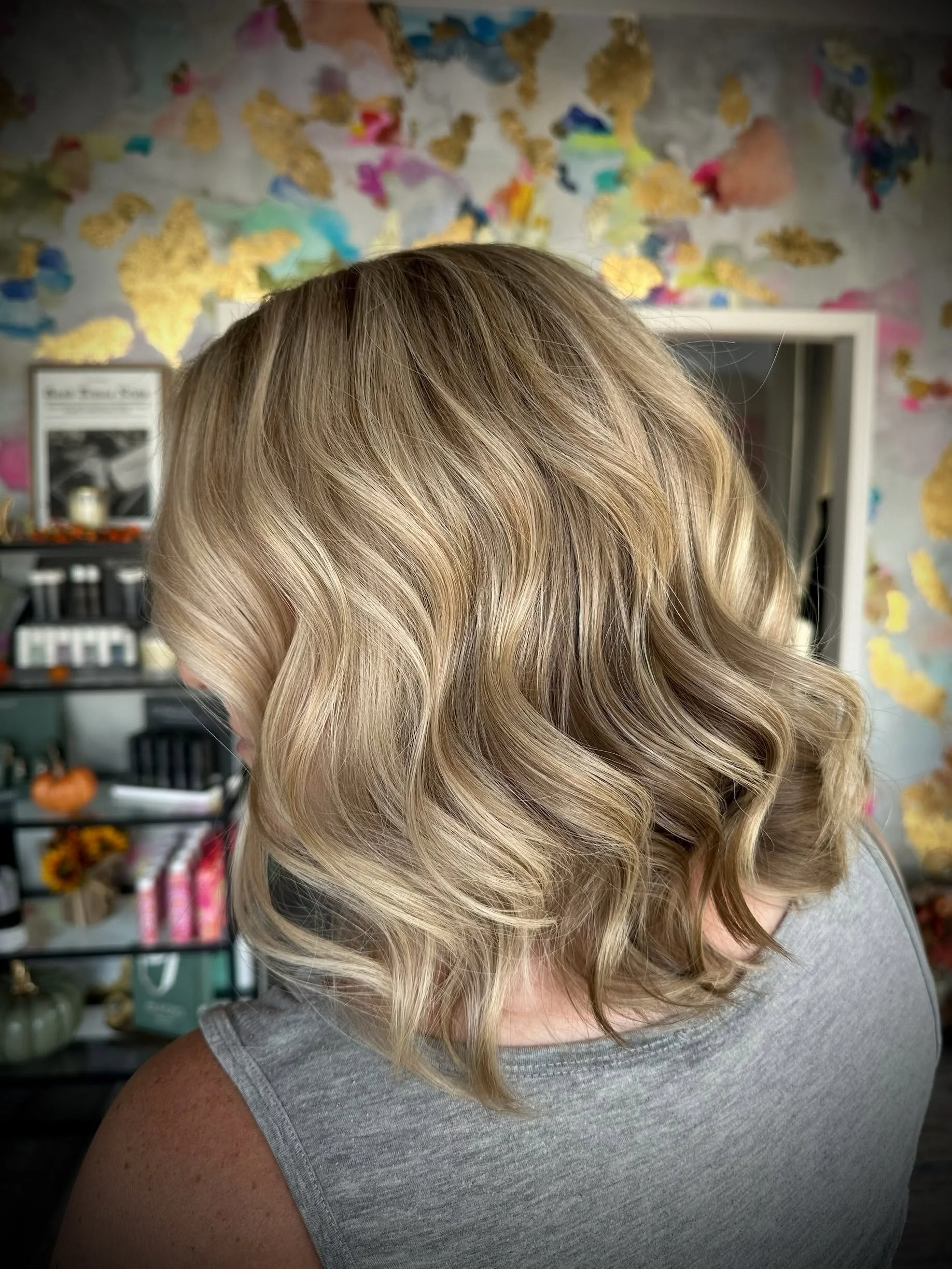 Close-up of a woman with shoulder-length blonde hair styled in loose waves, wearing a gray sleeveless top, with an interior background featuring colorful abstract wall art and shelves.