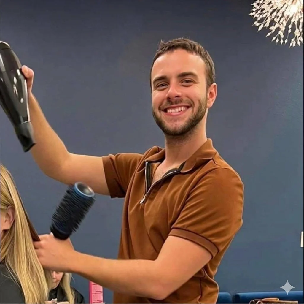 A smiling man blowing hair dry at a salon, dressed in a brown polo shirt, with a woman sitting in front of him.