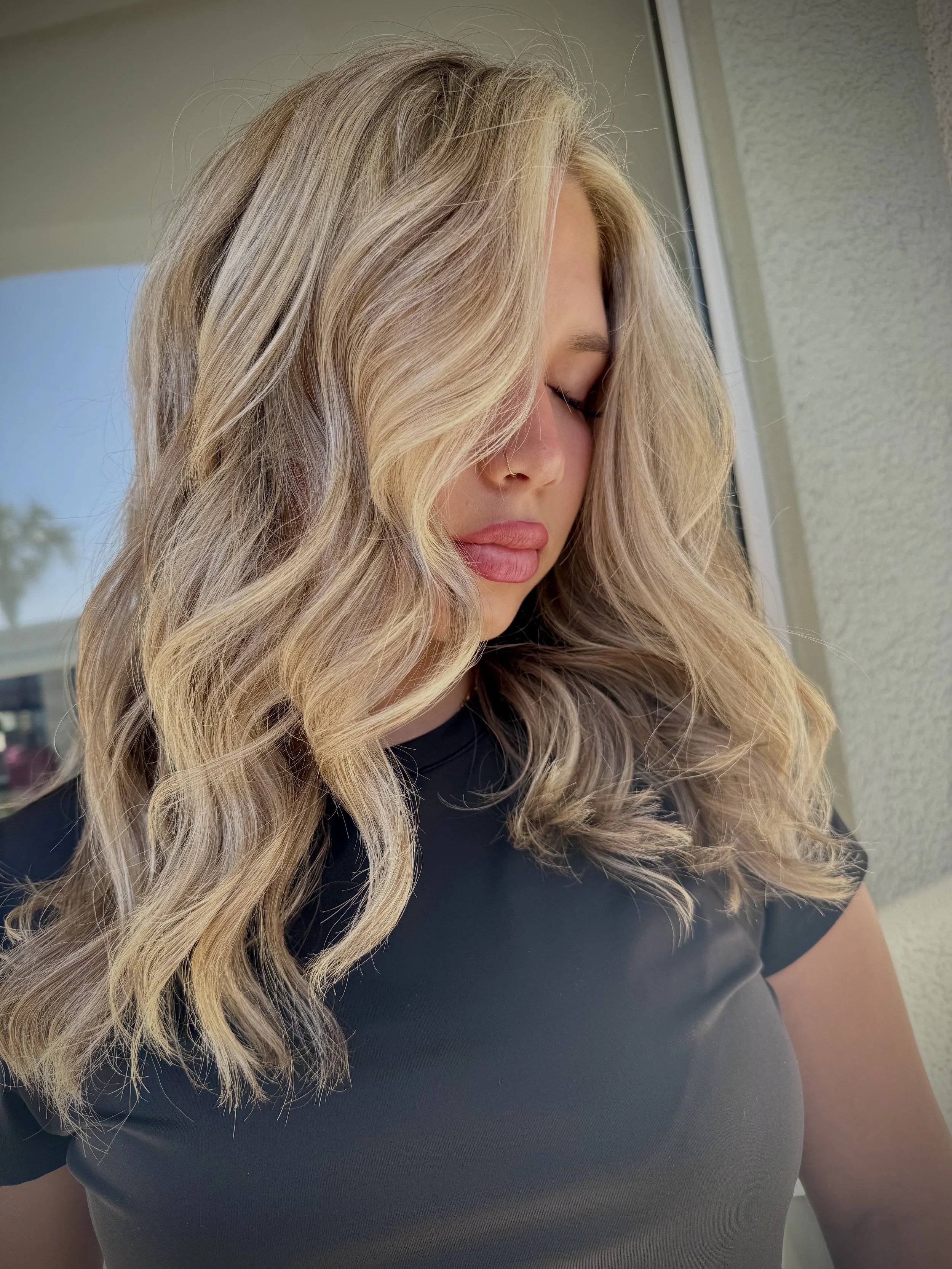 A close-up of a woman with long, wavy blonde hair cascading over her shoulder, wearing a black top, standing near a window with a blurred outdoor background.