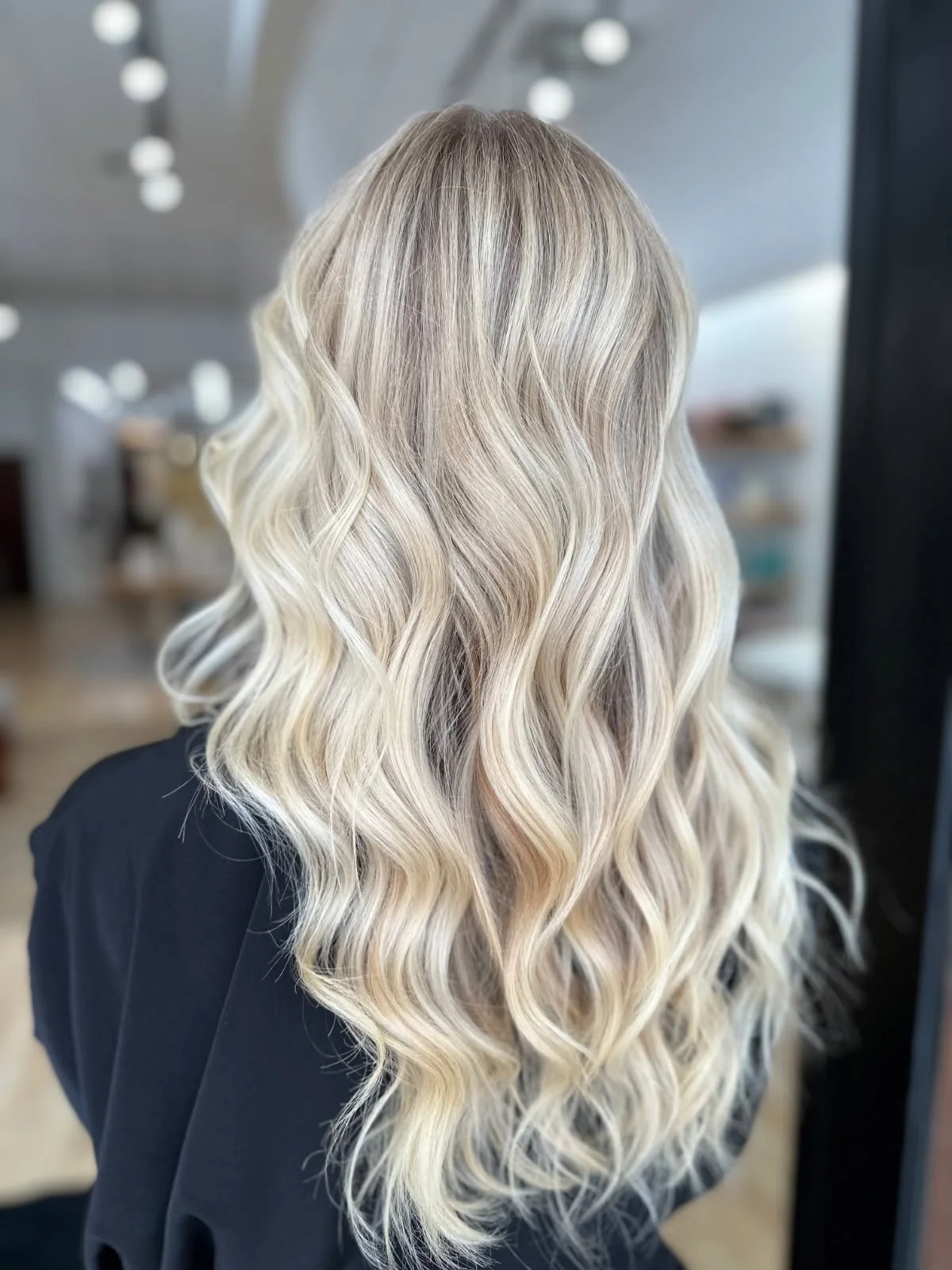 Back of a woman with long, wavy, blonde hair with darker roots, wearing a black top, in a salon or similar setting.