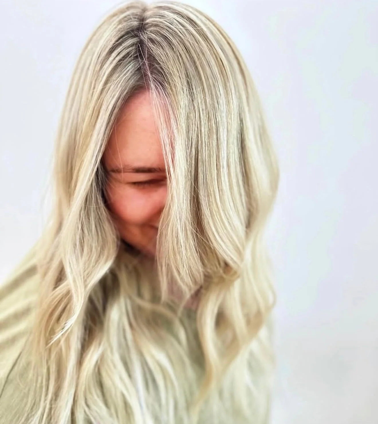 A woman with long, wavy blonde hair, smiling with eyes closed, captured against a plain white background.