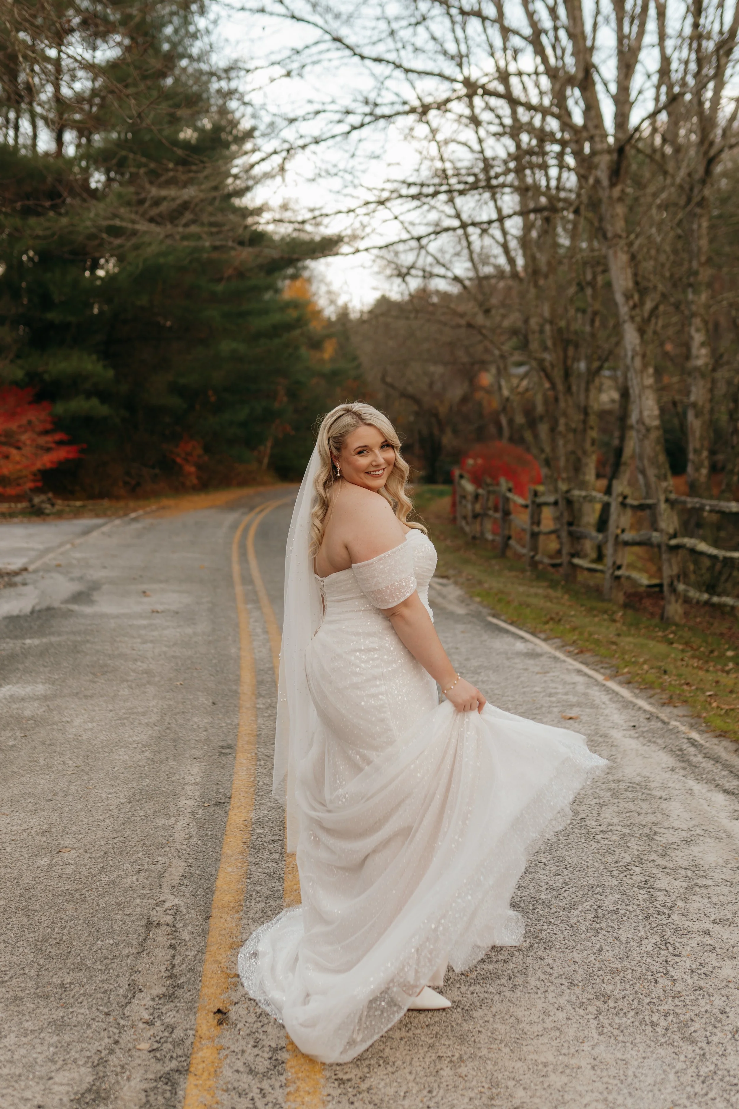 A smiling bride in a white wedding dress holding the hem of her dress, standing on an empty road surrounded by trees with autumn foliage.