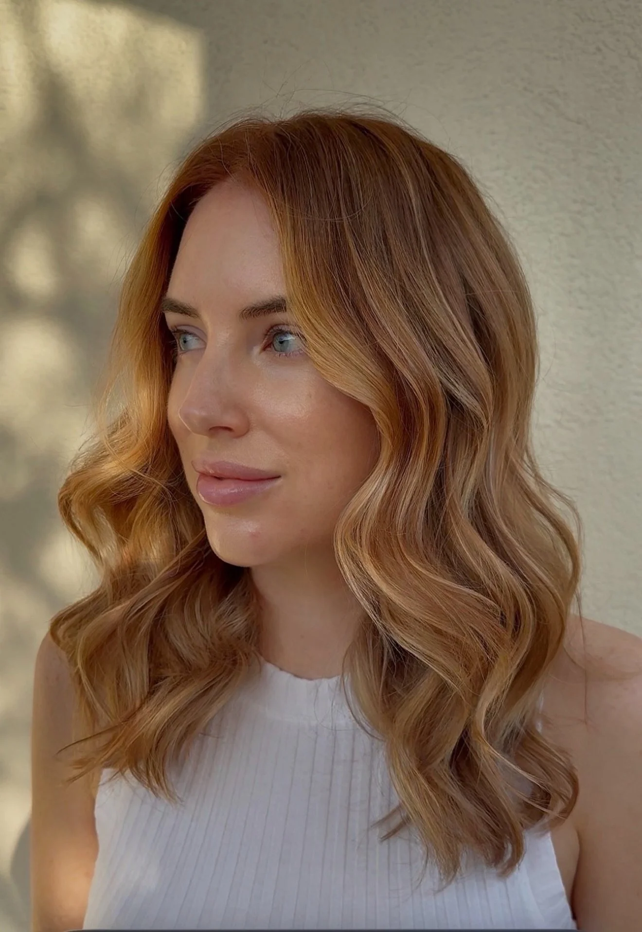 A woman with long, wavy red hair and fair skin looking slightly to the side, wearing a sleeveless white top.