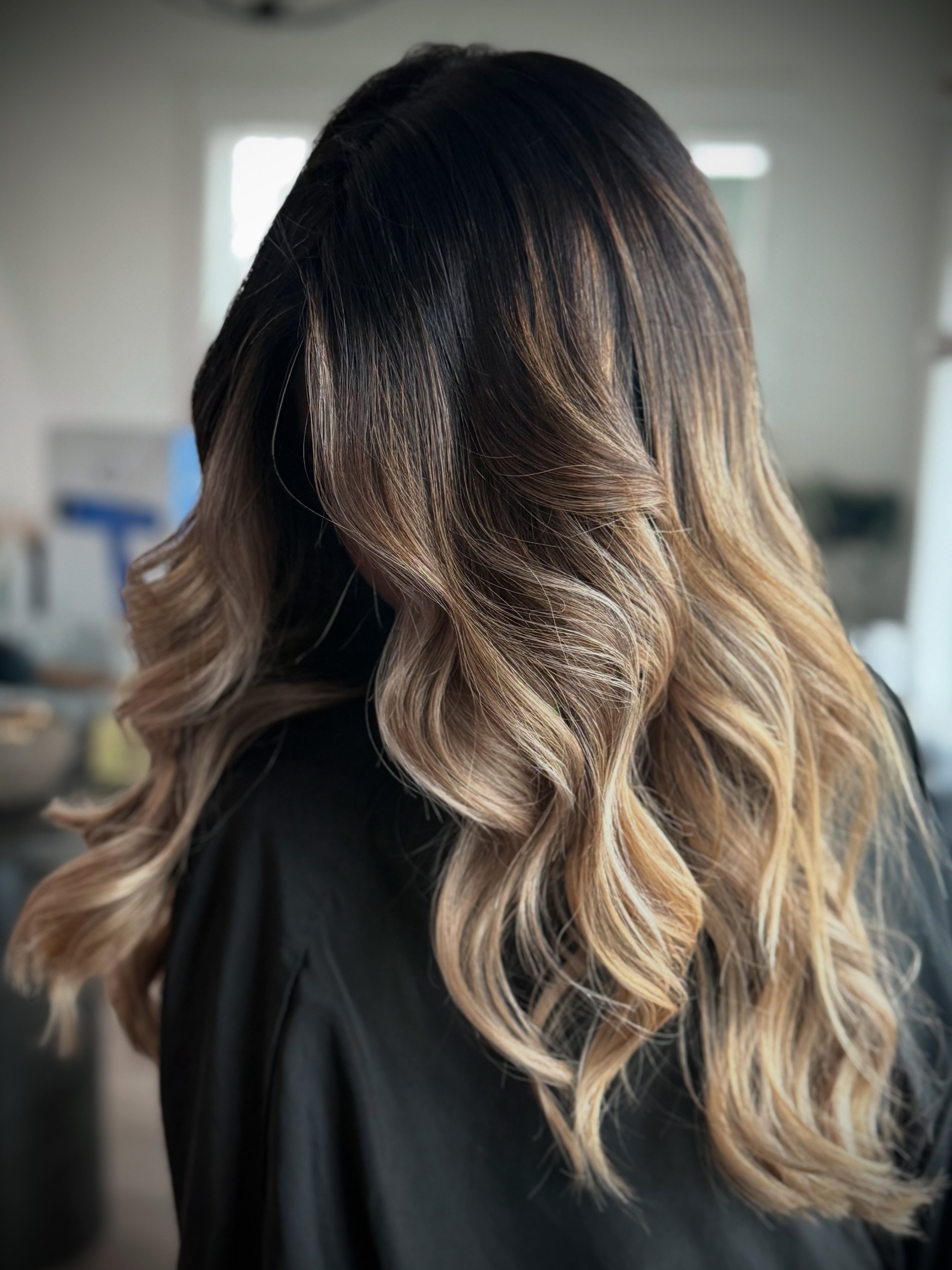 Woman with long, wavy hair styled with dark roots and blonde-ombré tips, wearing a black top, standing in a room with blurred background and natural light.