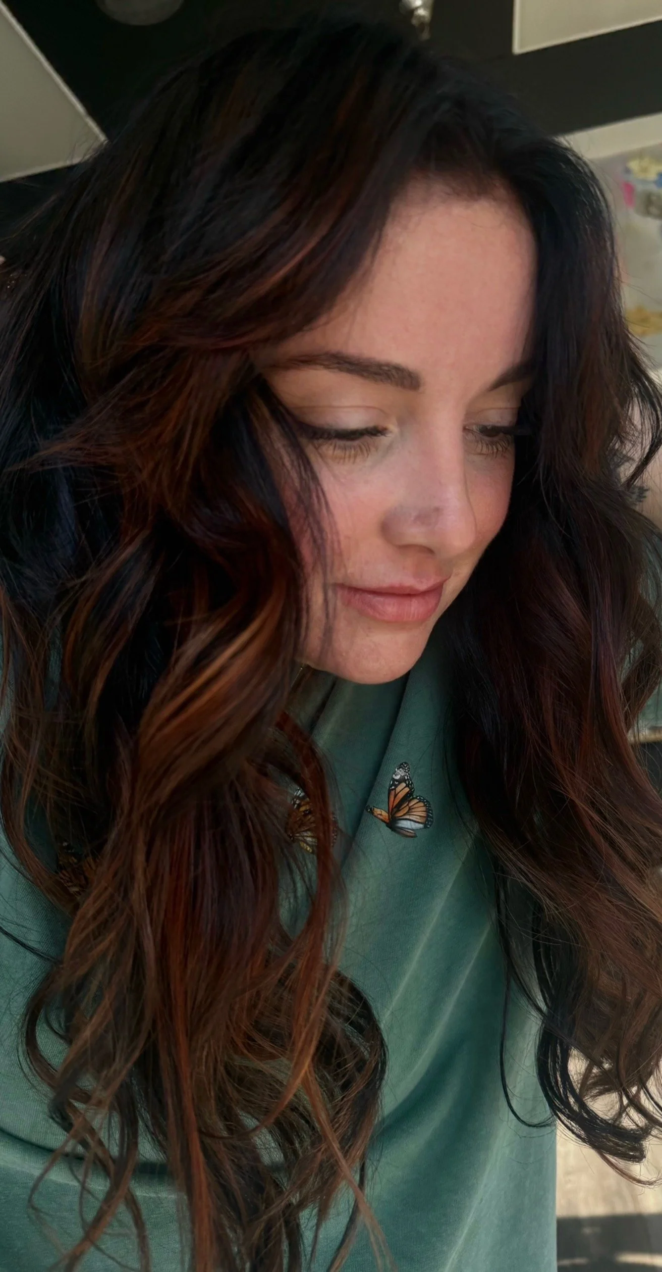 Close-up of a woman with long, wavy brunette hair with subtle highlights, wearing a green top with butterfly embroidery.