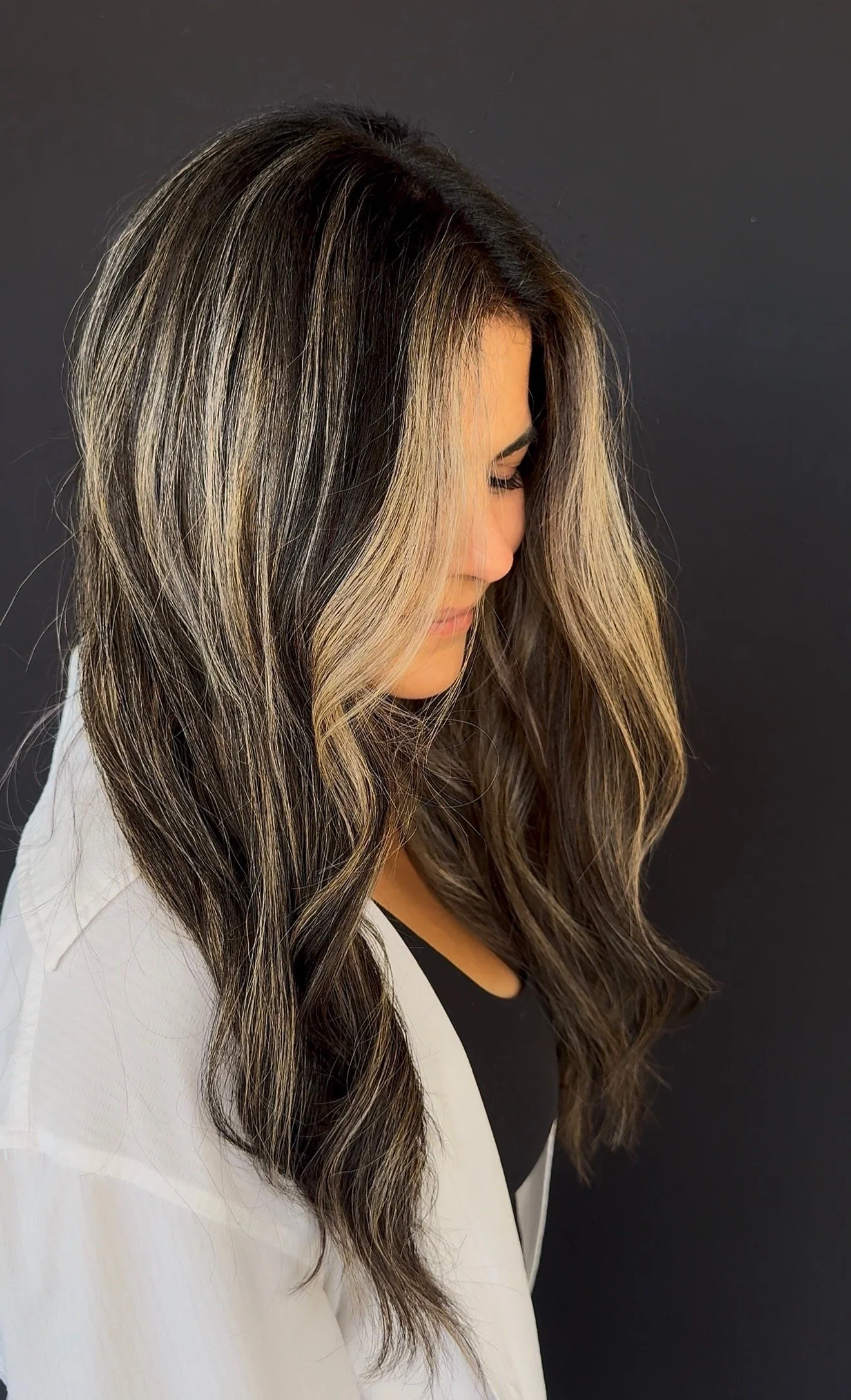 A woman with long, wavy hair that is dark at the roots and transitions to blonde highlights, facing downward with eyes closed against a plain dark background.