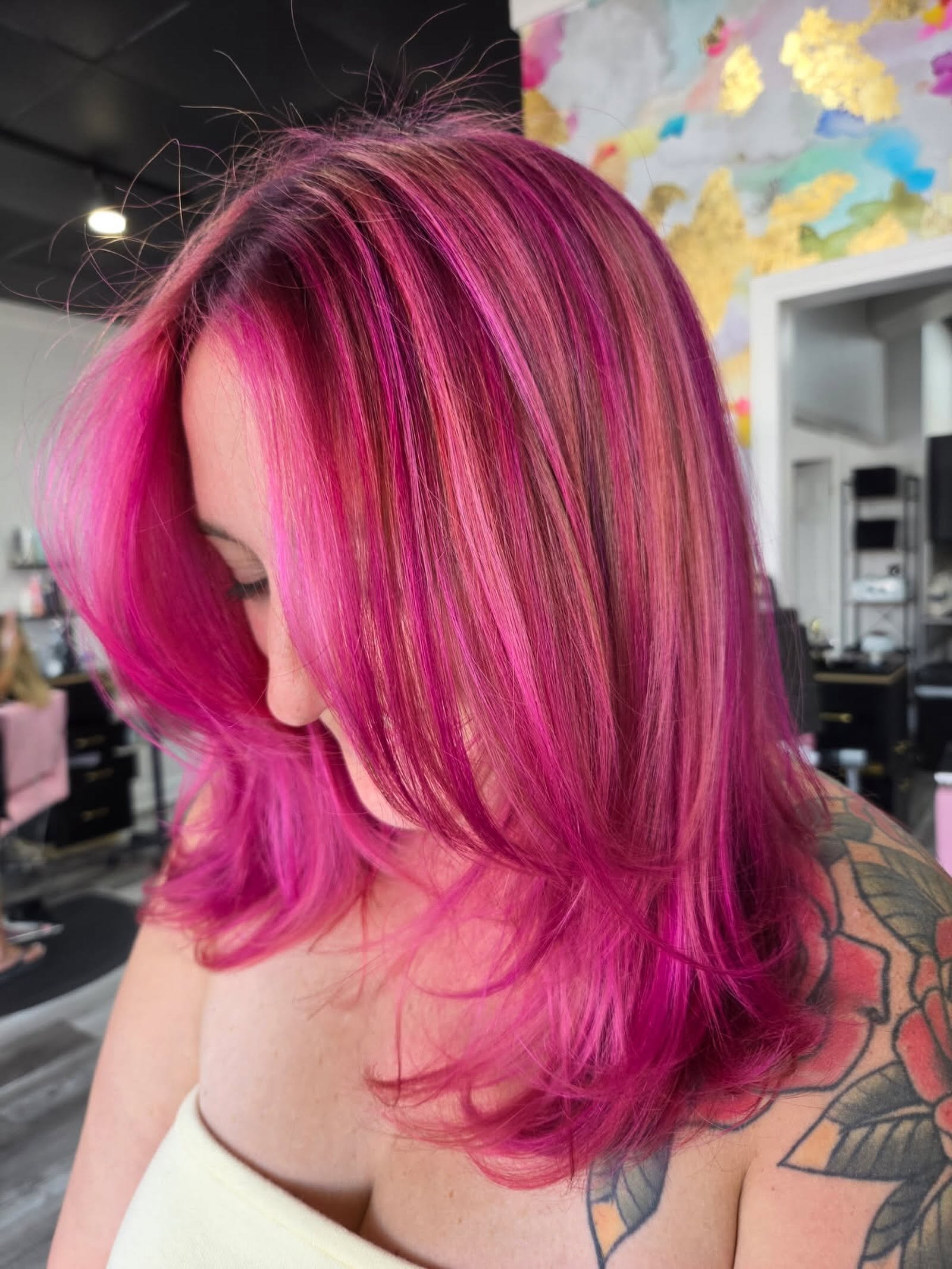 Close-up of a woman with shoulder-length vibrant pink hair with purple and lighter pink highlights, showing she has a tattoo on her shoulder.