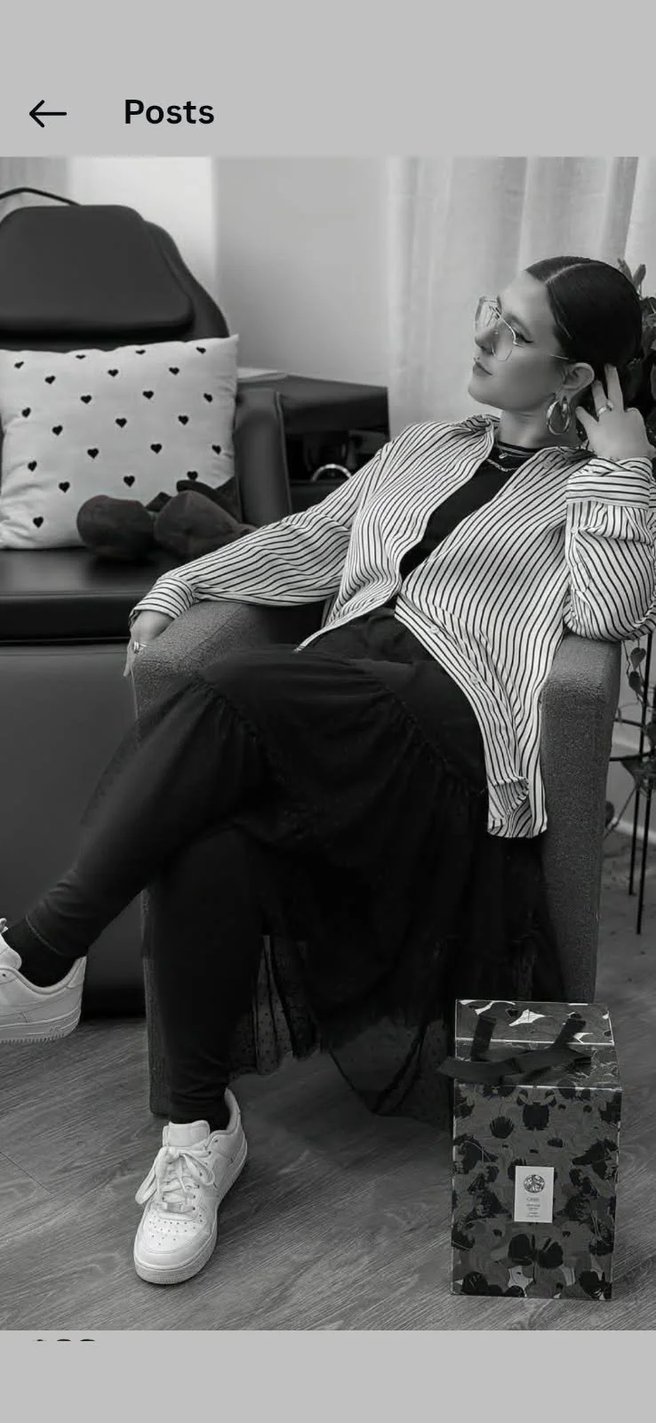 A woman sitting on an armchair, wearing glasses, a striped shirt over a black top, black skirt, and white sneakers. She has her hand behind her head and is looking to her left. There is a patterned gift bag on the floor next to her, and a pillow with heart shapes on a darker chair in the background.