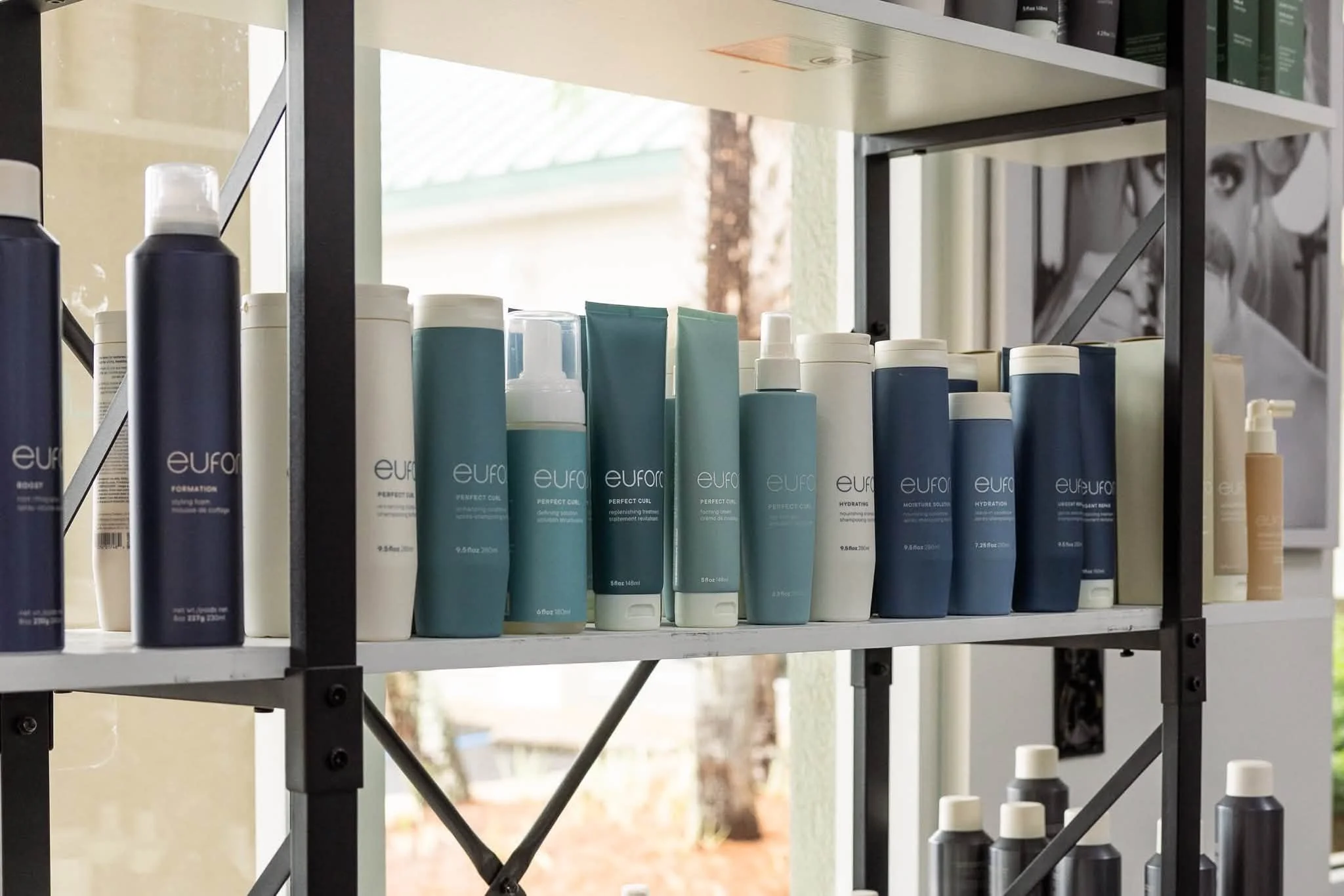 Shelf with various hair care products, including shampoos and conditioners in bottles of blue, white, and black, with a window in the background showing trees and a cloudy sky.