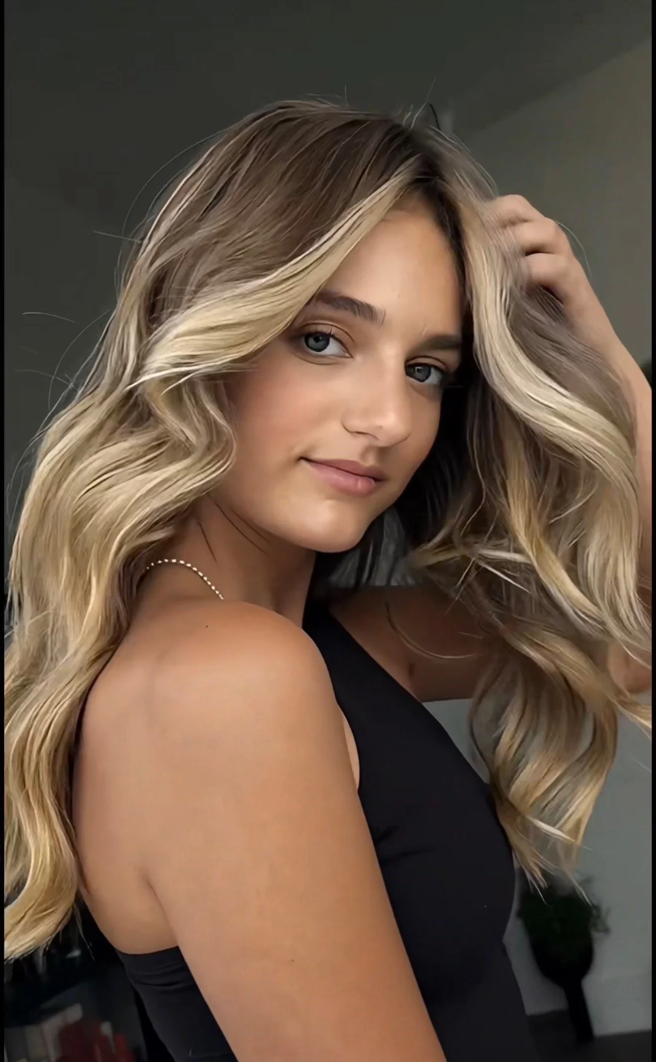 A woman with long blonde hair styled in loose waves, wearing a black sleeveless top, with her hand touching her hair, looking at the camera against a gray background.