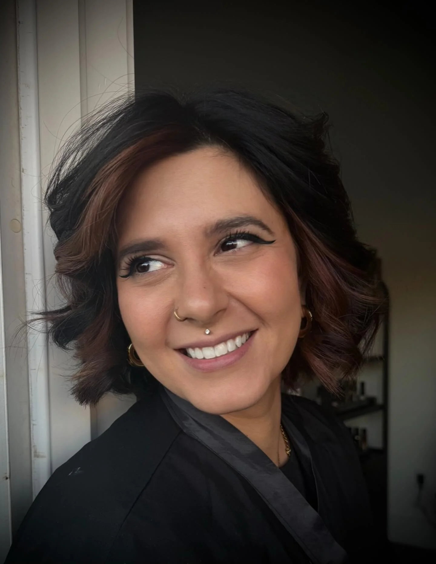 A woman with dark wavy hair, wearing makeup, earrings, and a black outfit, smiling and looking to her left, in front of a door and a dark background.