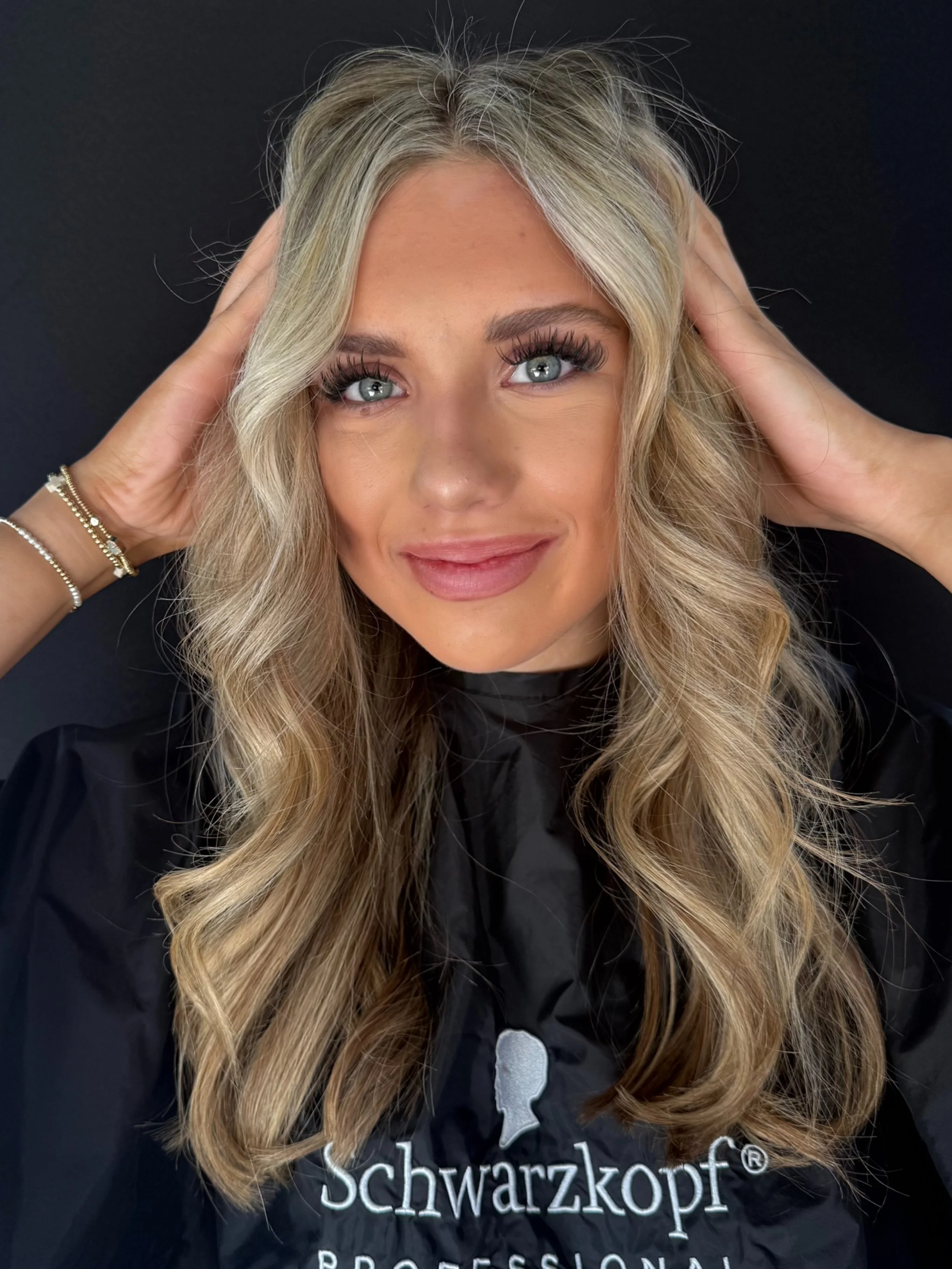 Close-up of a woman with long blonde wavy hair, light skin, and blue eyes, smiling softly, touching her temples. She is wearing a black shirt with the word 'schwarzkopf' and some additional text in white.