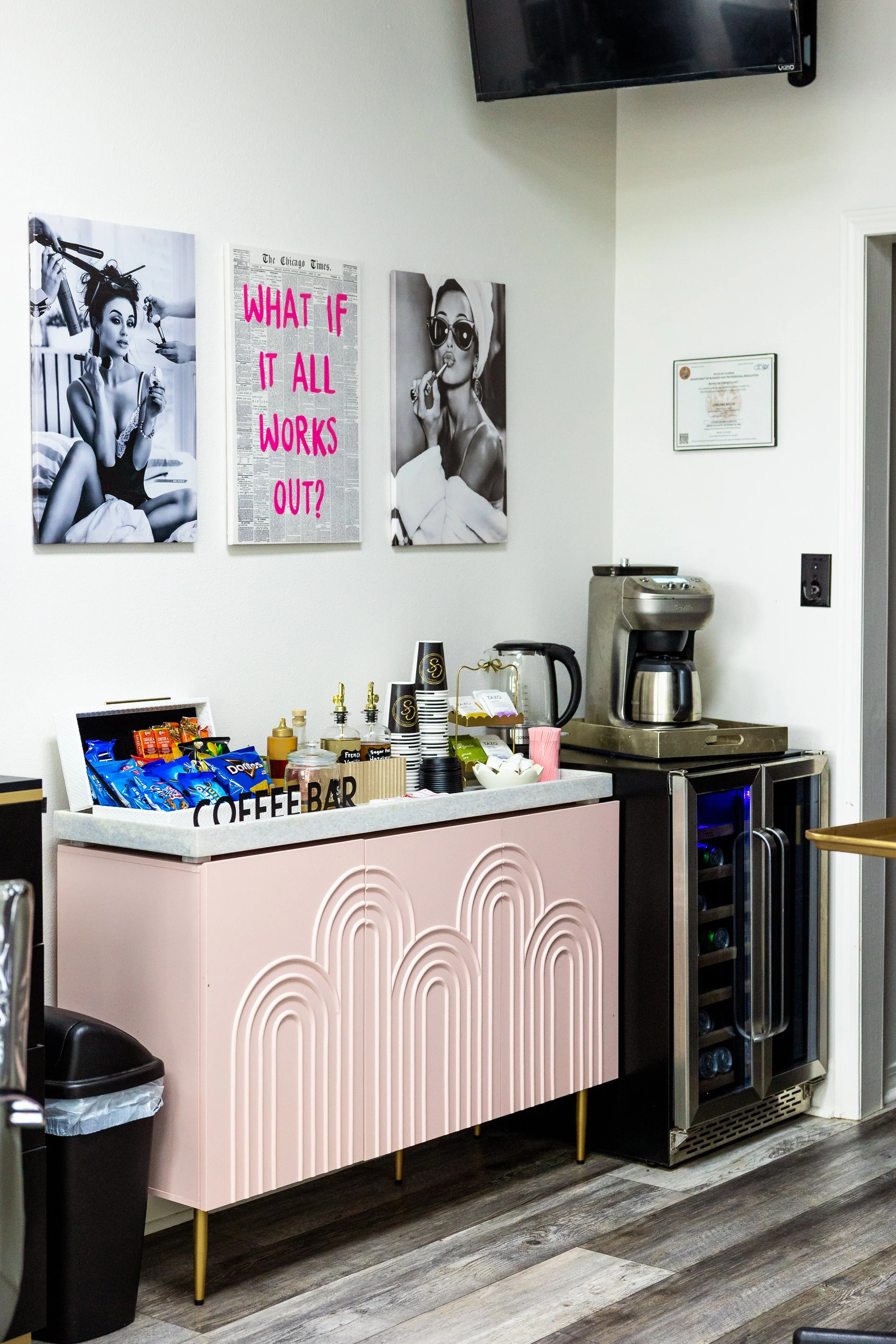 A coffee bar setup with a pink decorated cabinet, snack bags, coffee cups, and a coffee machine. On the wall, there are black-and-white photos of women and a newspaper with pink text reading 'What if it all works out?'.