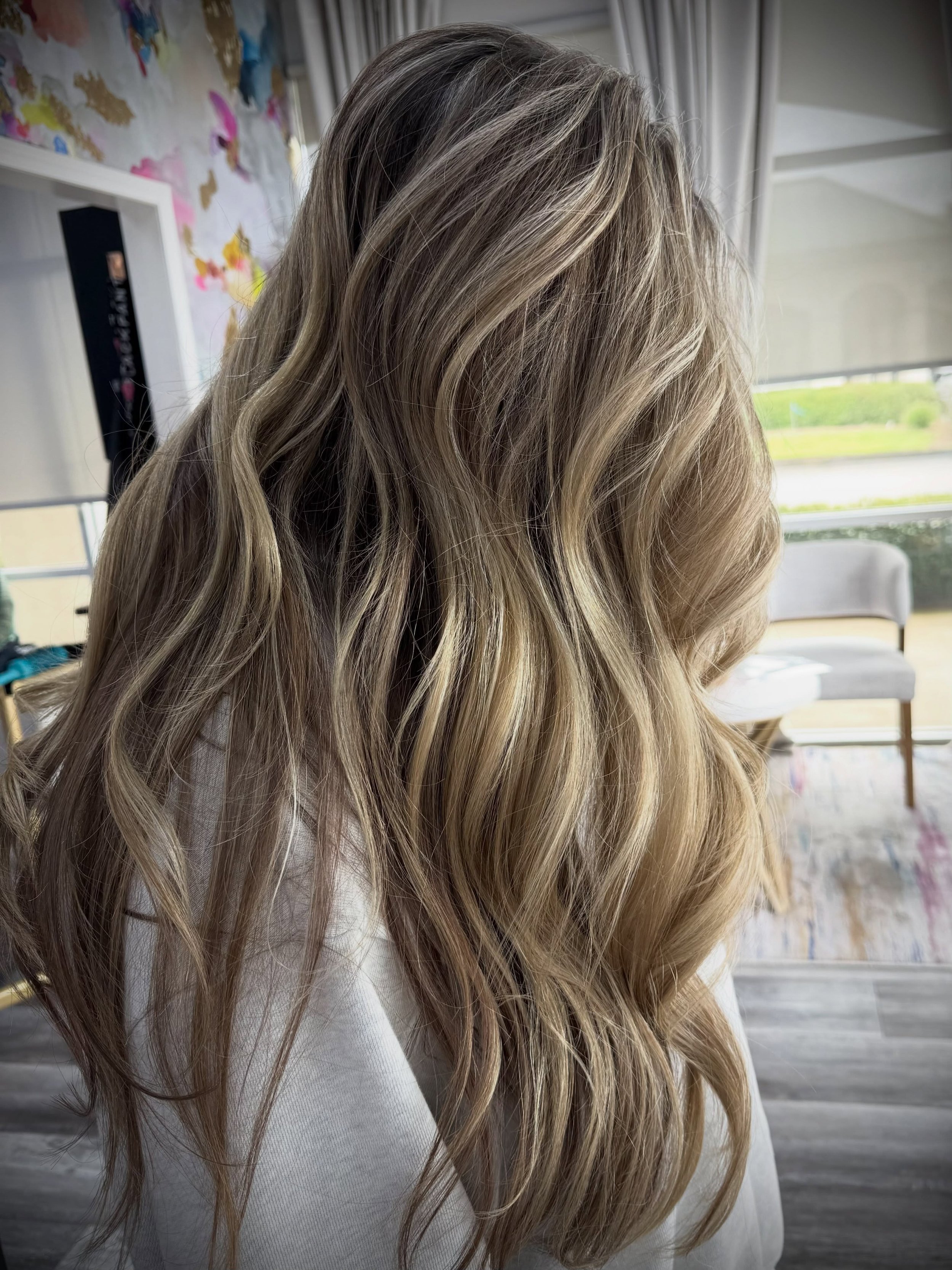 Close-up of blonde wavy hair, with an indoor background including a chair, curtains, and a window.