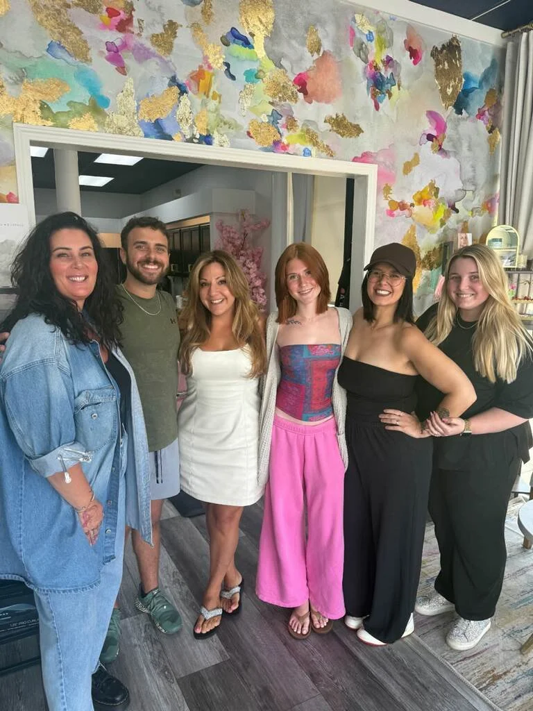 Six women smiling and posing together inside a colorful, artistic, and vibrant room with floral and abstract designs on the wall.