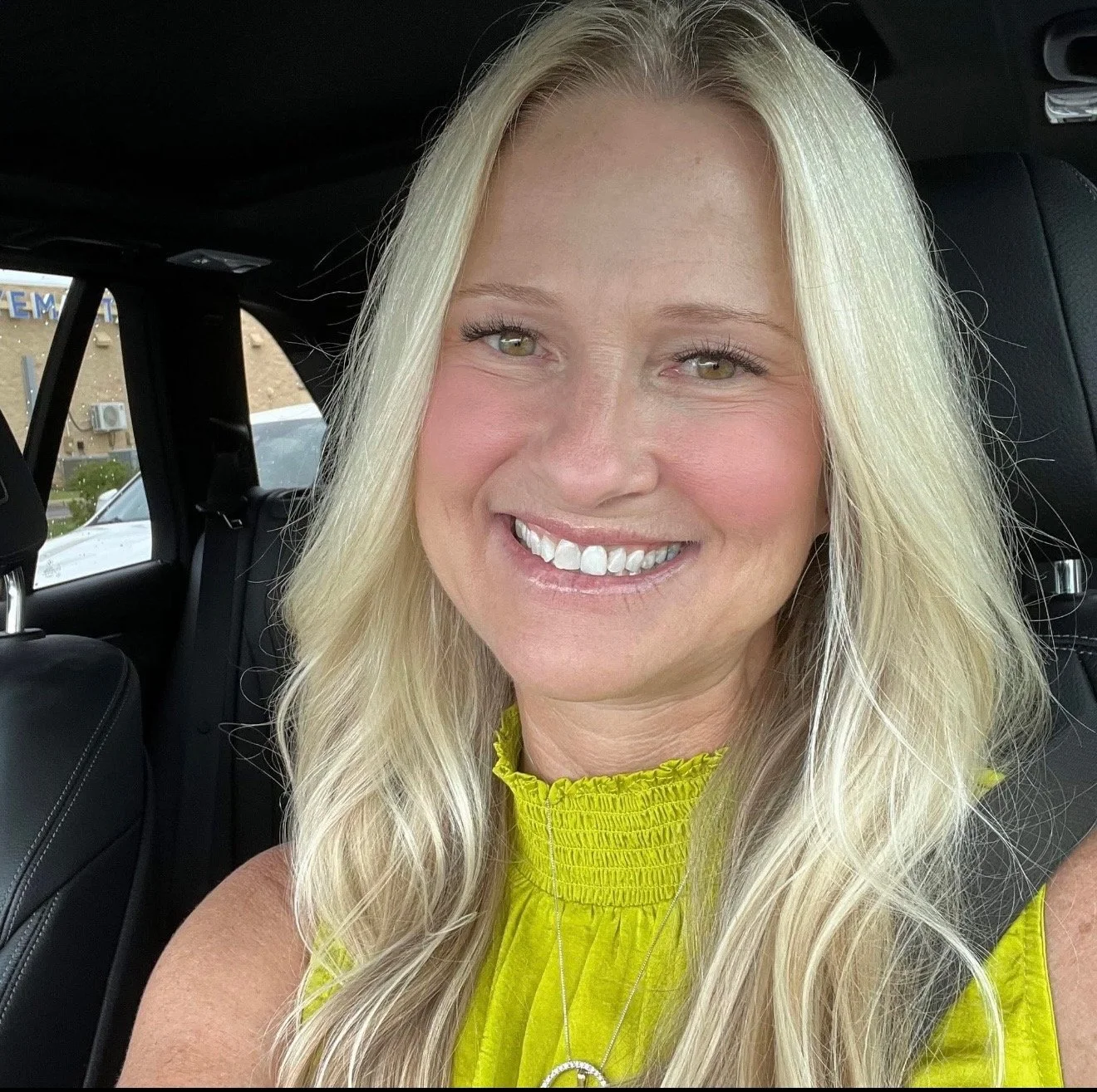 A woman with long blonde hair smiling in a car, wearing a yellow sleeveless top.