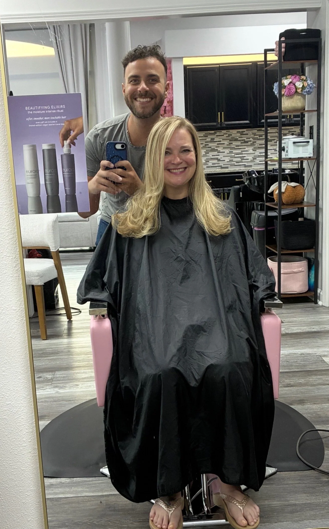 A woman with blonde hair sitting in a hair salon chair, wearing a black salon cape, smiling for a mirror selfie. A hairstylist with curly hair stands behind her, taking the photo with a smartphone. The salon has shelves with flowers, towels, and salo