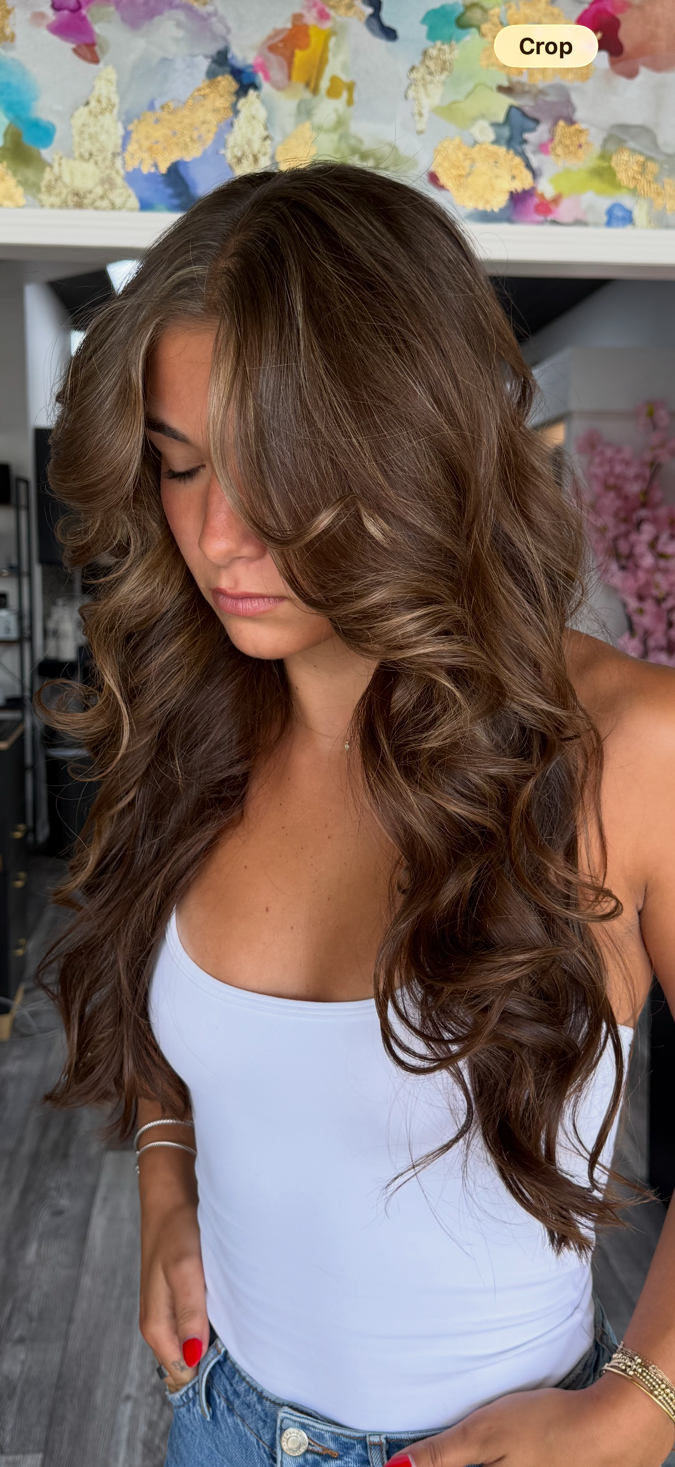 A woman with long, curly brown hair, wearing a white tank top, gold jewelry, and painted red nails, standing indoors with a colorful abstract painting on the wall behind her.