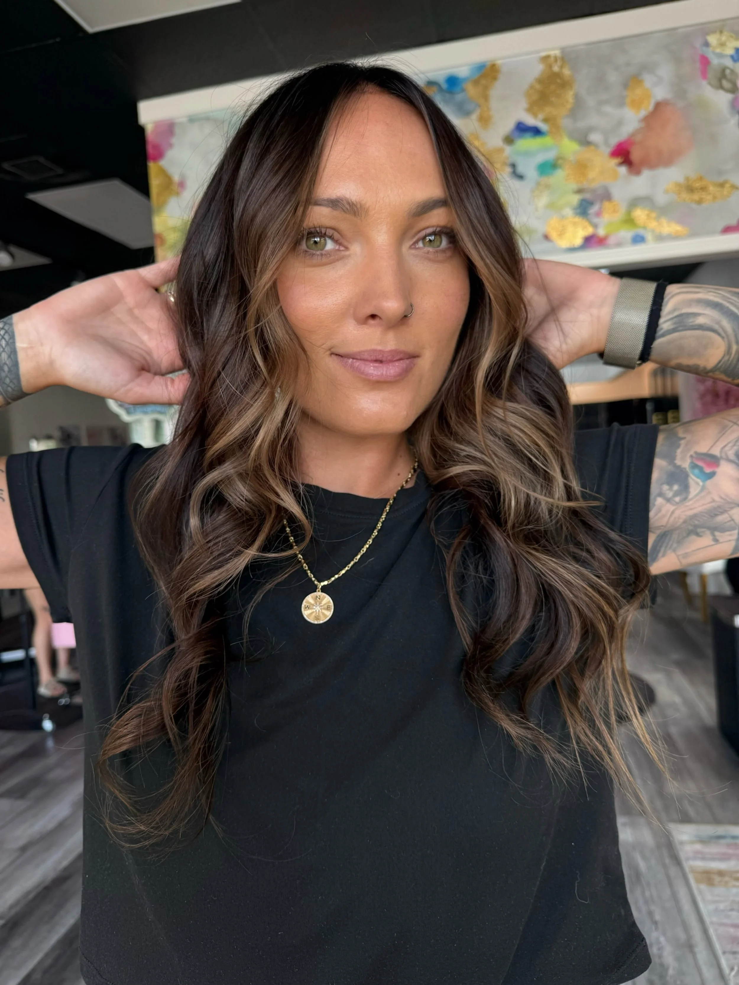 A woman with long wavy hair and light green eyes, wearing a black shirt, gold necklace, and a nose ring, standing in a studio with her hands behind her head, with colorful abstract artwork in the background.