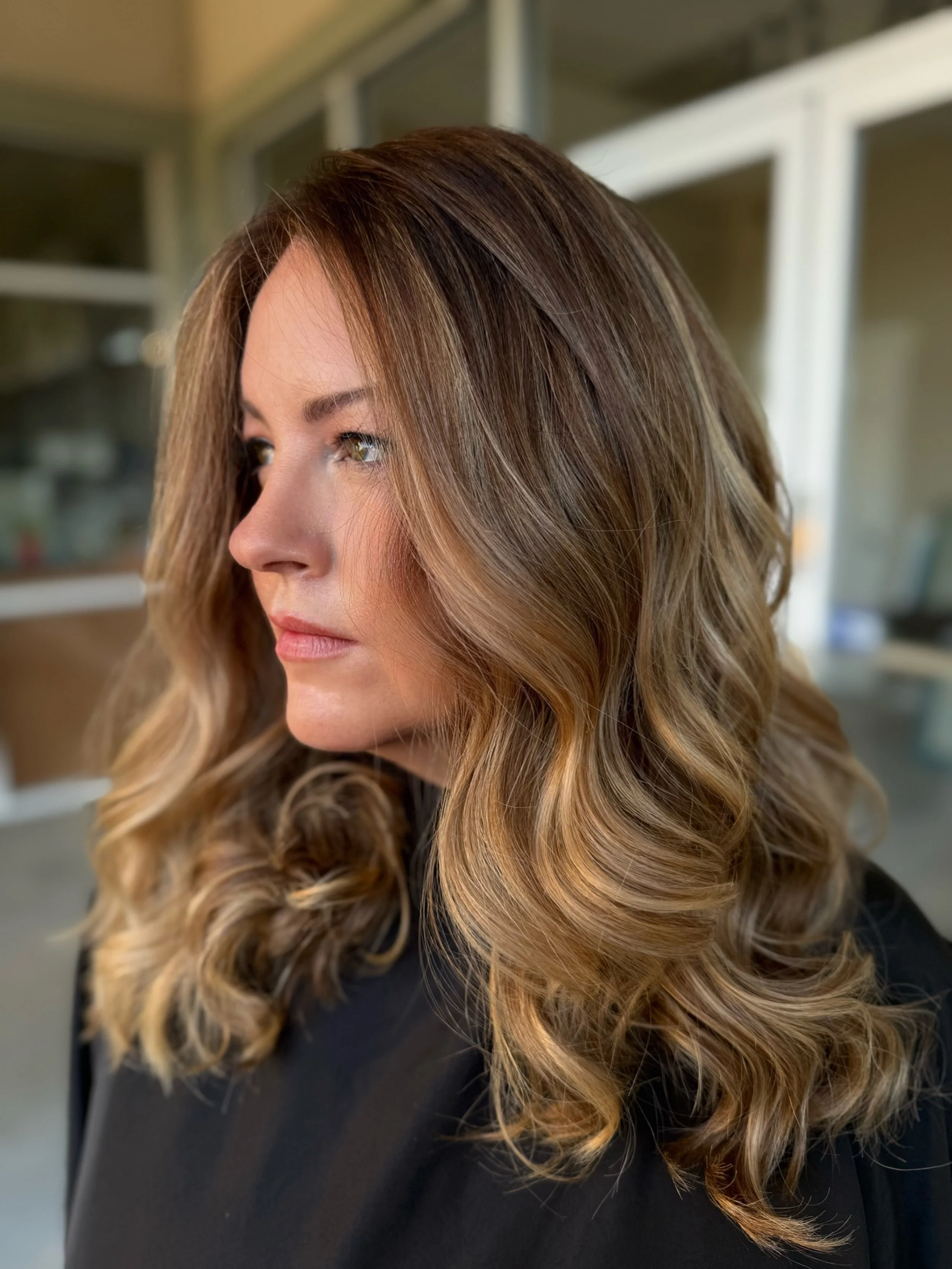 Close-up of a woman with wavy, ombre hair ranging from dark blonde to light caramel, wearing a black top, standing indoors with a blurred background.