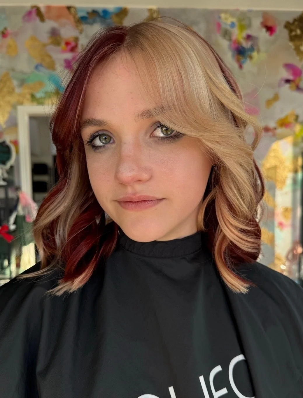 A woman with shoulder-length wavy hair, partially blonde with reddish-brown streaks, and light makeup, looking at the camera, in a hair salon with a colorful butterfly-themed background.