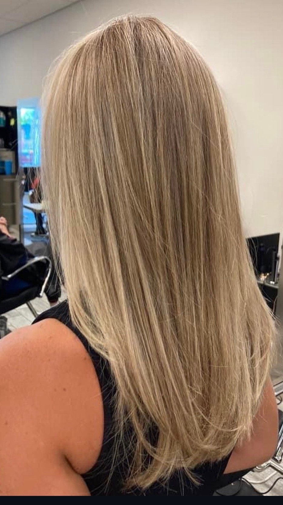 Blonde woman with straight hair sitting in a salon or waiting area, with a vending machine or refreshment area in the background.
