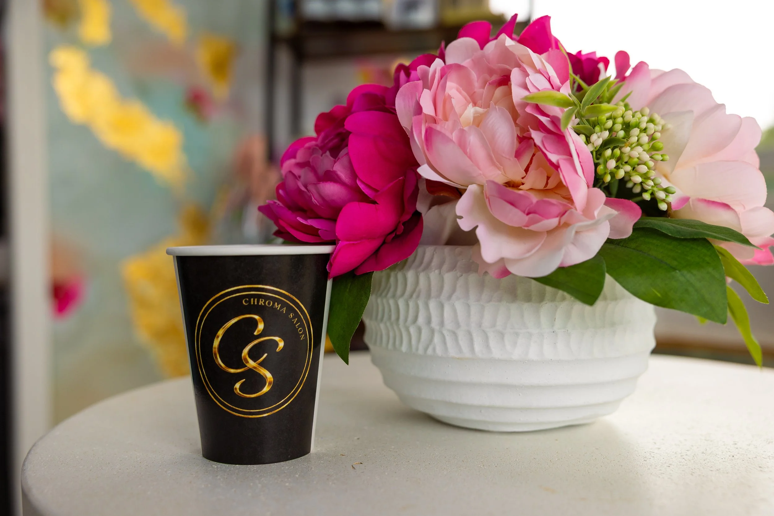 Close-up of pink and white flowers in a white vase with green leaves, positioned next to a black coffee cup with gold lettering.