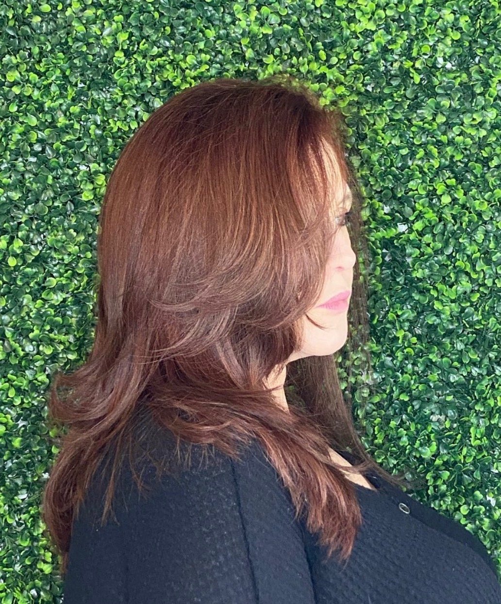 Side profile of a woman with long, wavy reddish-brown hair, standing against a lush green leafy backdrop.