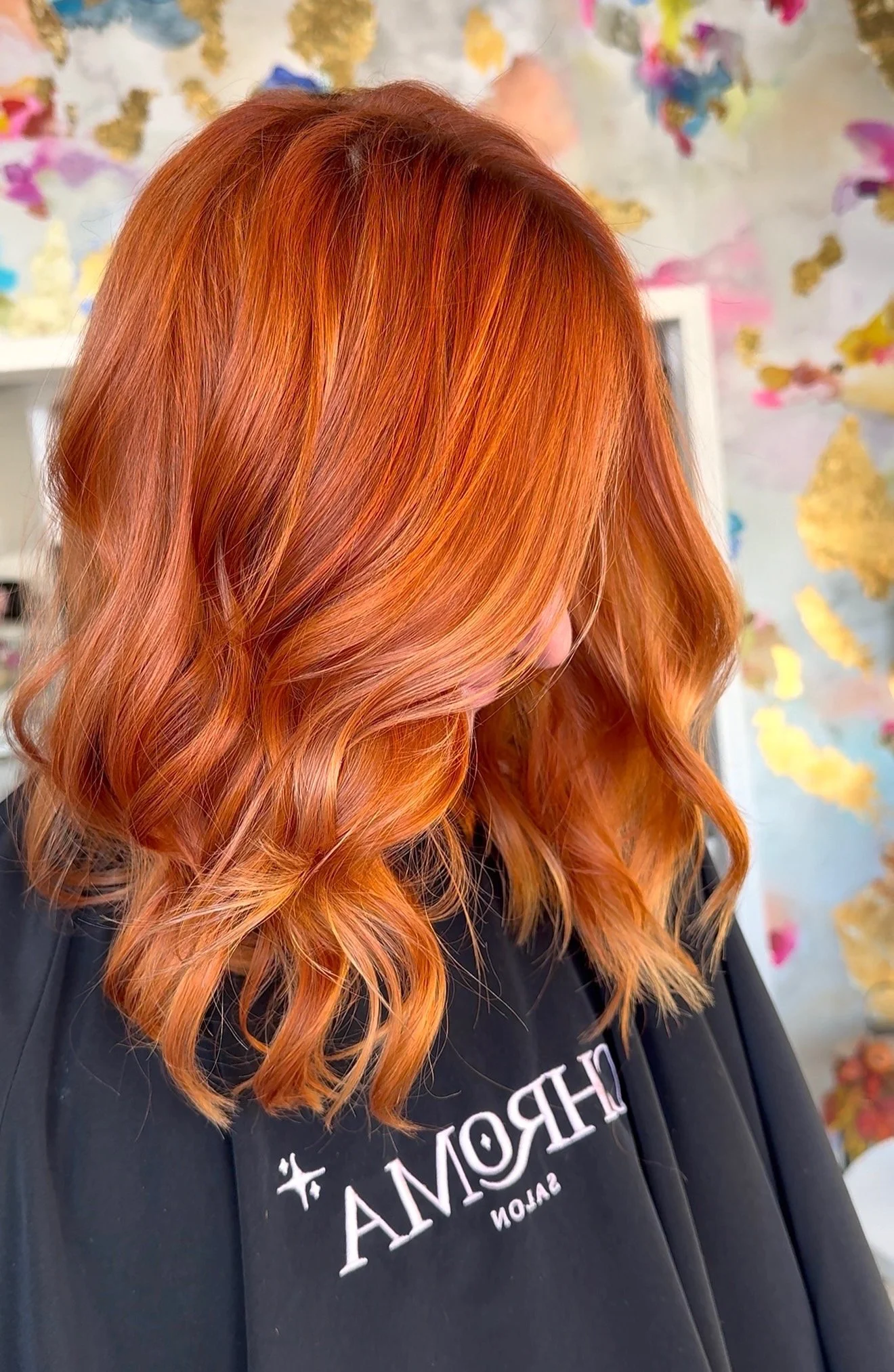 Close-up of a woman with vibrant red-orange hair styled in loose curls, wearing a black salon apron with white text, standing in front of a colorful, abstract background.