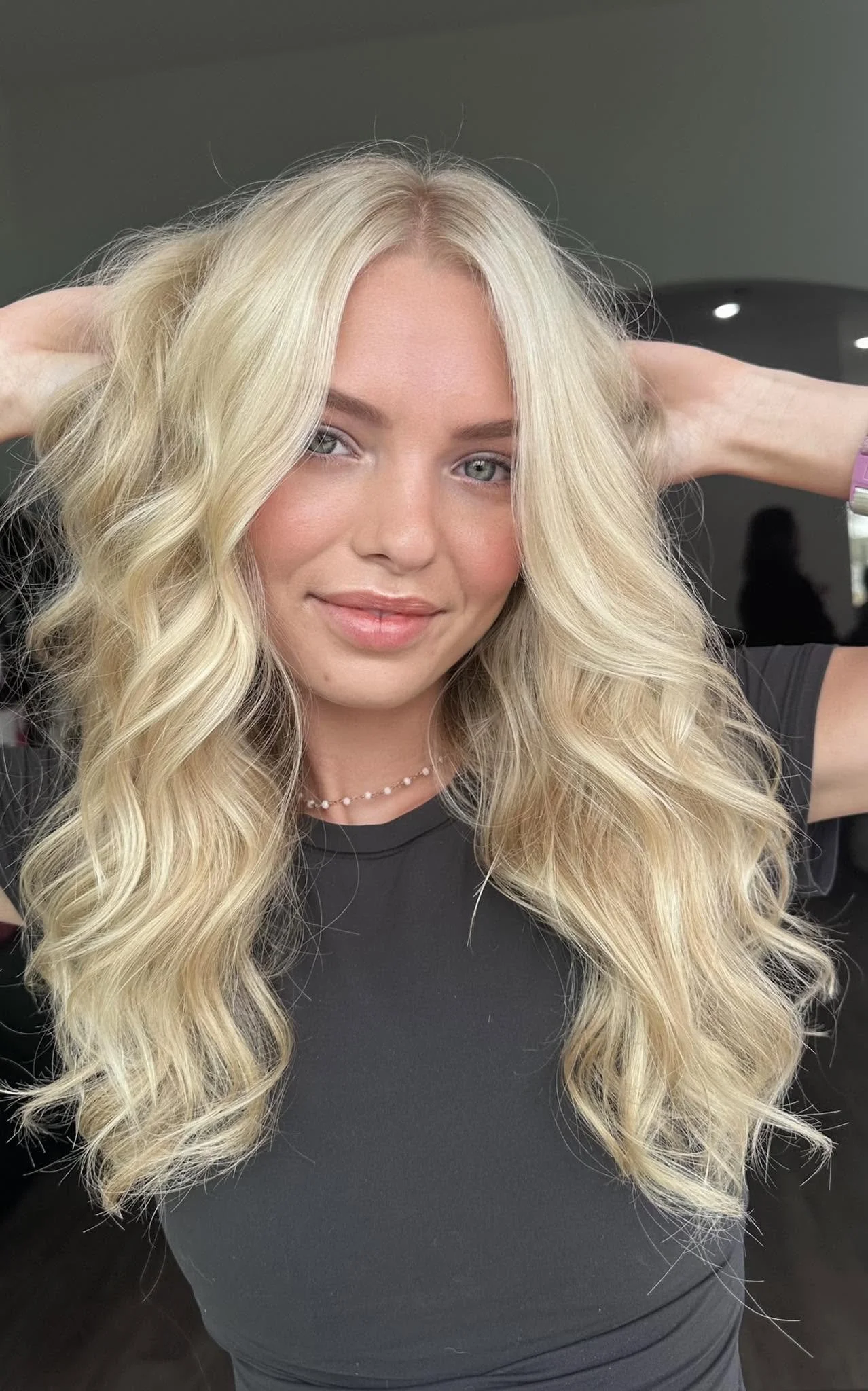 A young woman with long, wavy blonde hair and fair skin, smiling slightly and looking into the camera. She is wearing a black shirt, a delicate pearl necklace, and a purple wristband or watch.