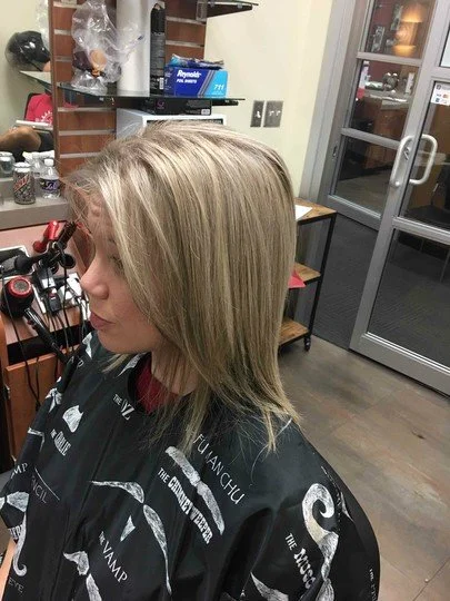 A woman with blonde hair sitting in a salon chair in front of a mirror, wearing a black cape with white text and graphics, in a salon with shelves, items, and a door in the background.