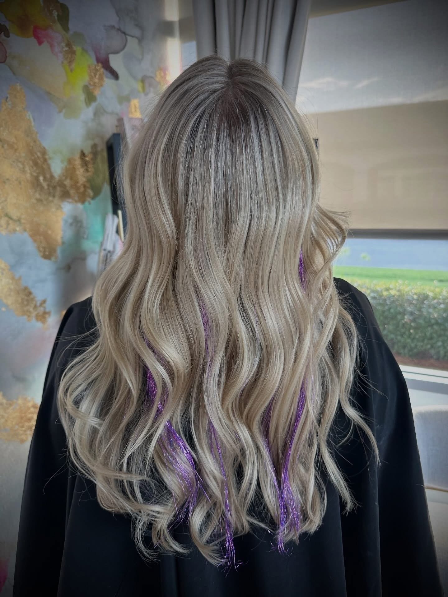 Back view of a person with long, wavy blonde hair, showing purple streaks, wearing a black cape, standing in front of a window with a view of greenery and a curtain, with a world map in the background.