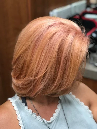 A woman with short, ombre hair with shades of peach and blonde, wearing a light blue top with lace trim and a necklace, in a hair salon.
