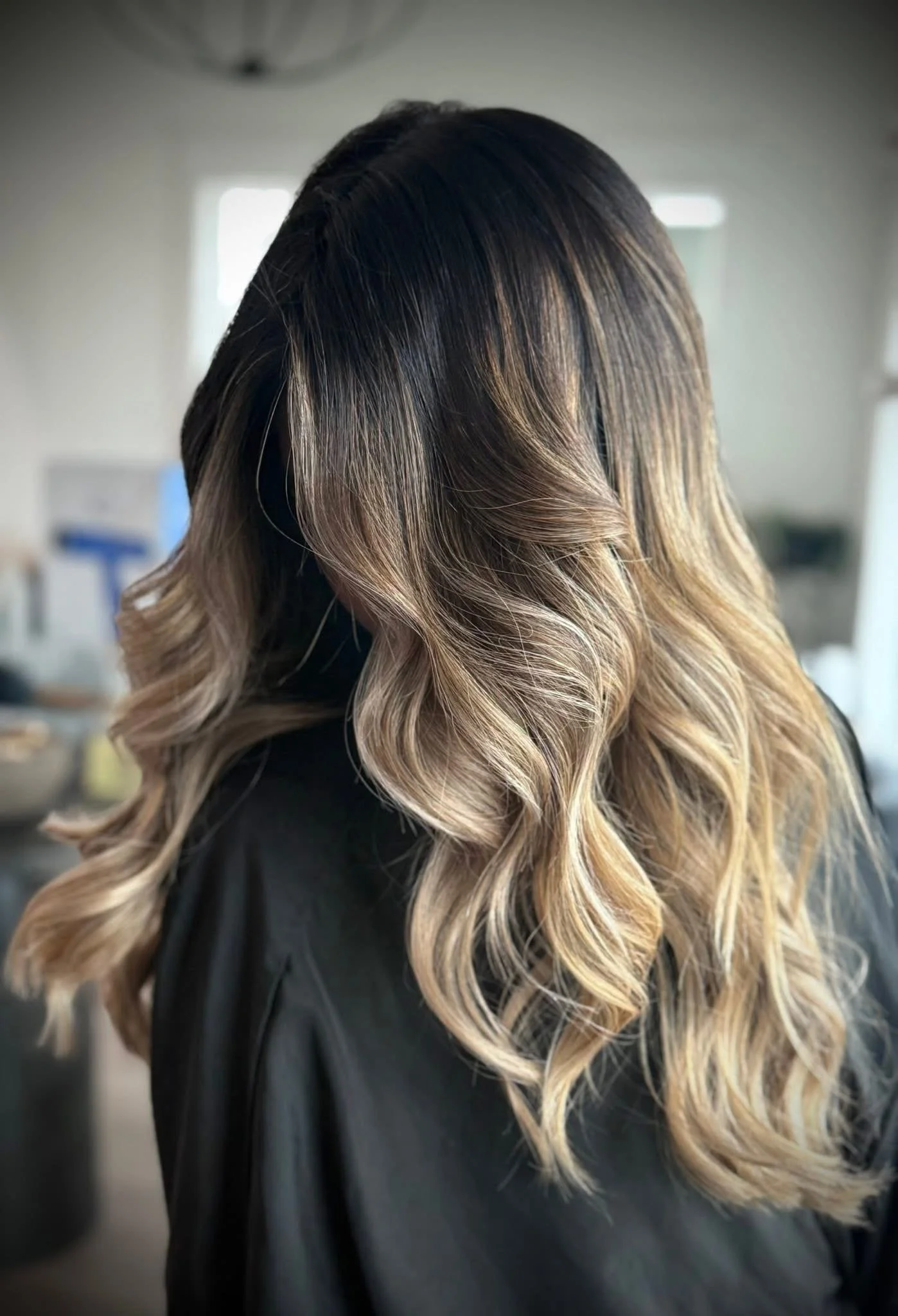 Close-up of a woman with dark roots and blonde, wavy hair, in a salon or indoor setting.