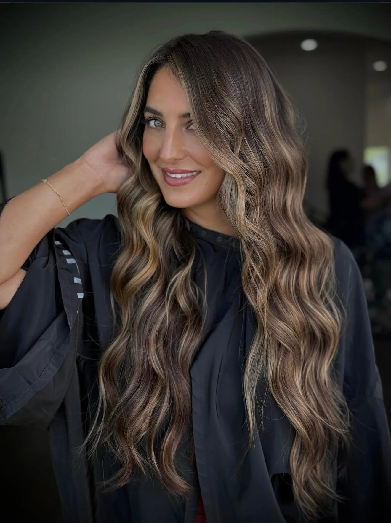 Woman with long wavy hair getting a haircut at a salon, smiling, with background figures of other people.