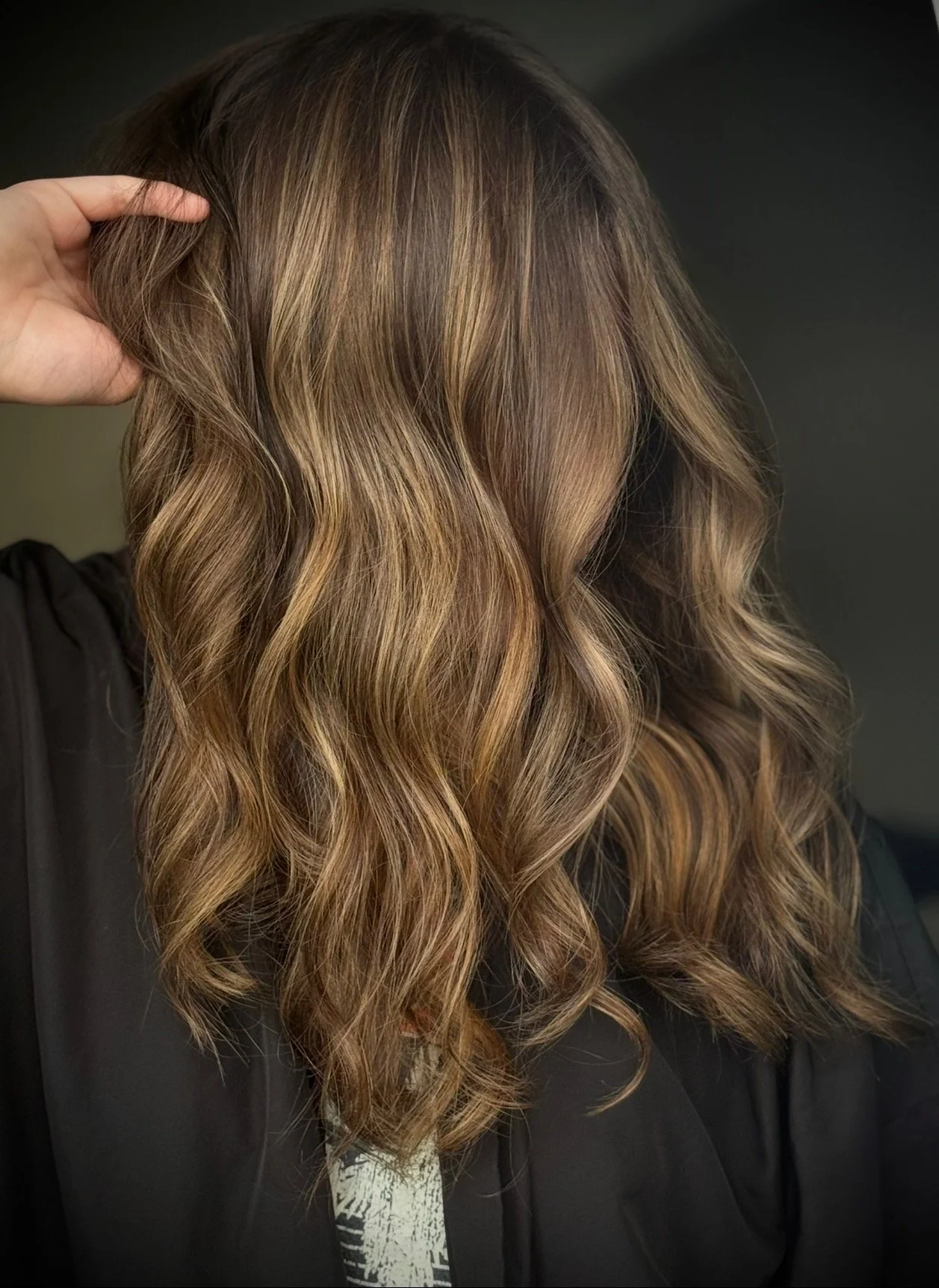 Hair stylist's hand touching long, wavy, light brown hair with darker lowlights standing against a dark background.