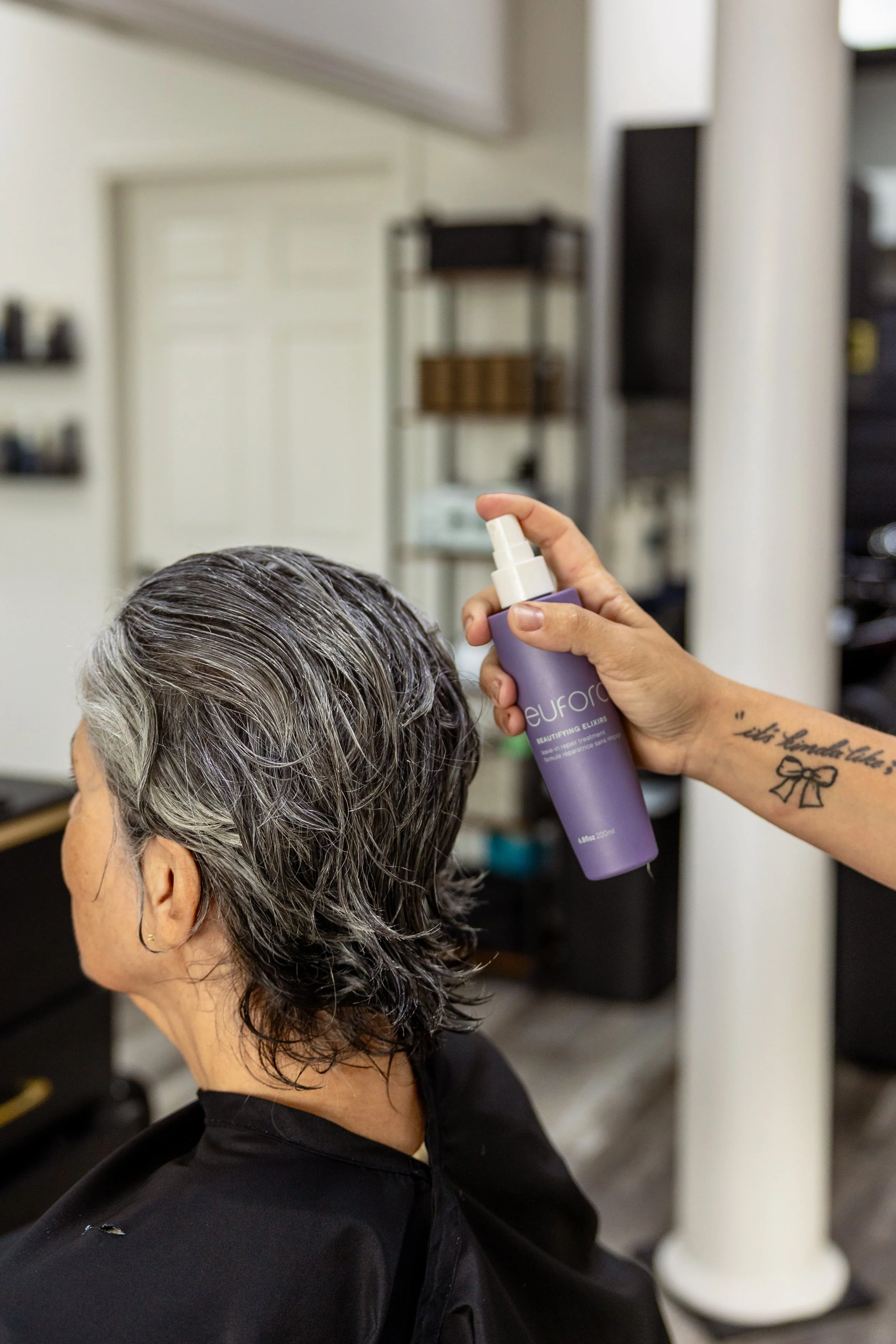 A person with short, gray hair getting their hair sprayed with a purple bottle labeled 'EUFORA' in a salon or barbershop.