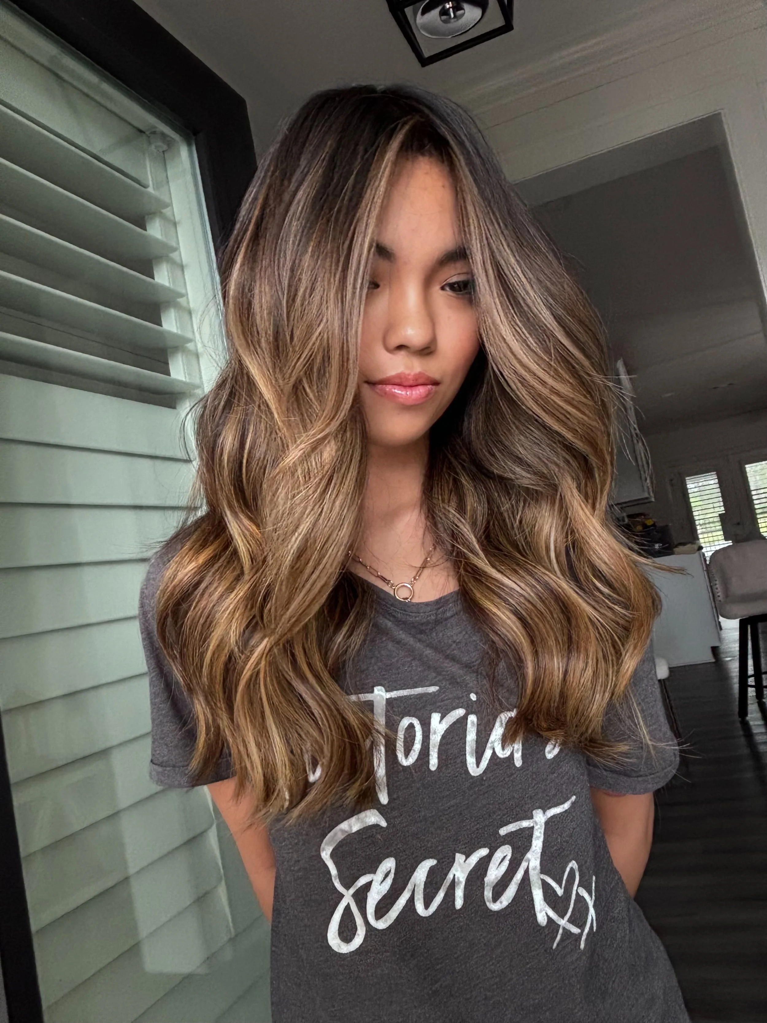 A woman with long, wavy hair with highlights standing indoors near a green house wall and a window. She is wearing a dark gray T-shirt with white cursive writing and a small necklace. The photo is focused on her face and upper body.