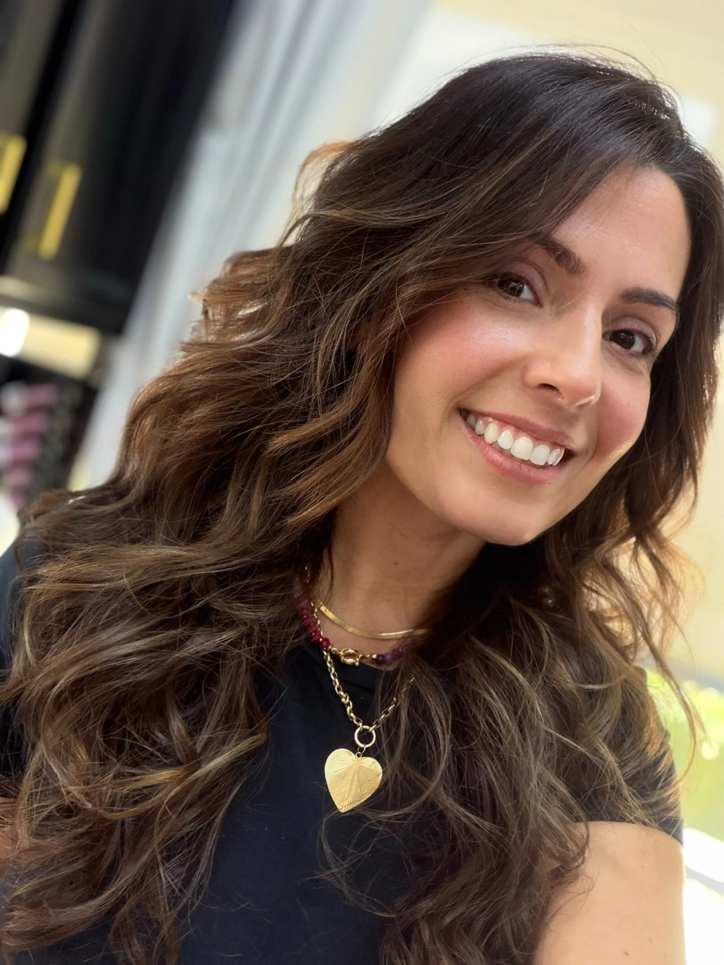 Close-up of a smiling woman with long, wavy brown hair, wearing multiple gold necklaces with decorative pendants, in an indoor setting.
