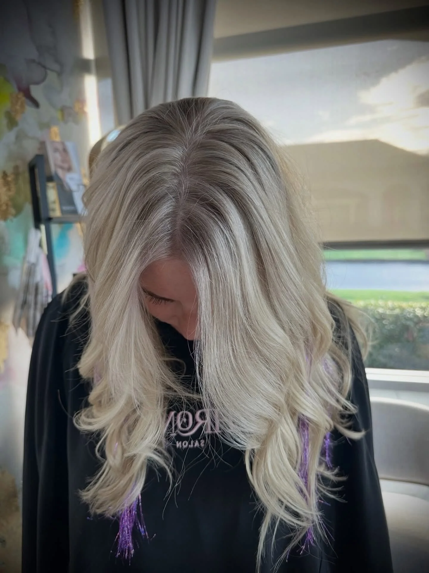 A woman with long, platinum blonde hair with gray roots, looking downward, inside a room with a window showing a cloudy sky. She's wearing a black shirt with a graphic design.