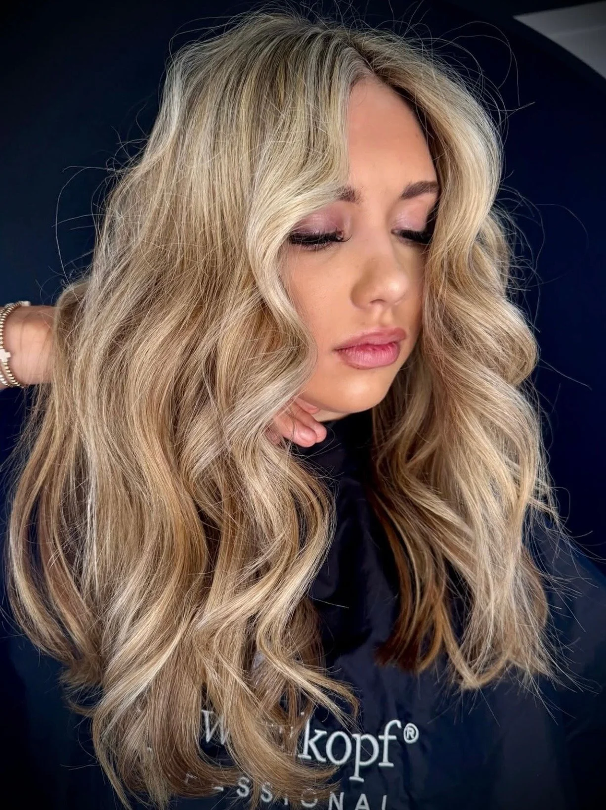 Close-up of a woman with long, wavy blonde hair, looking down with closed eyes, wearing a dark top and a silver bracelet.