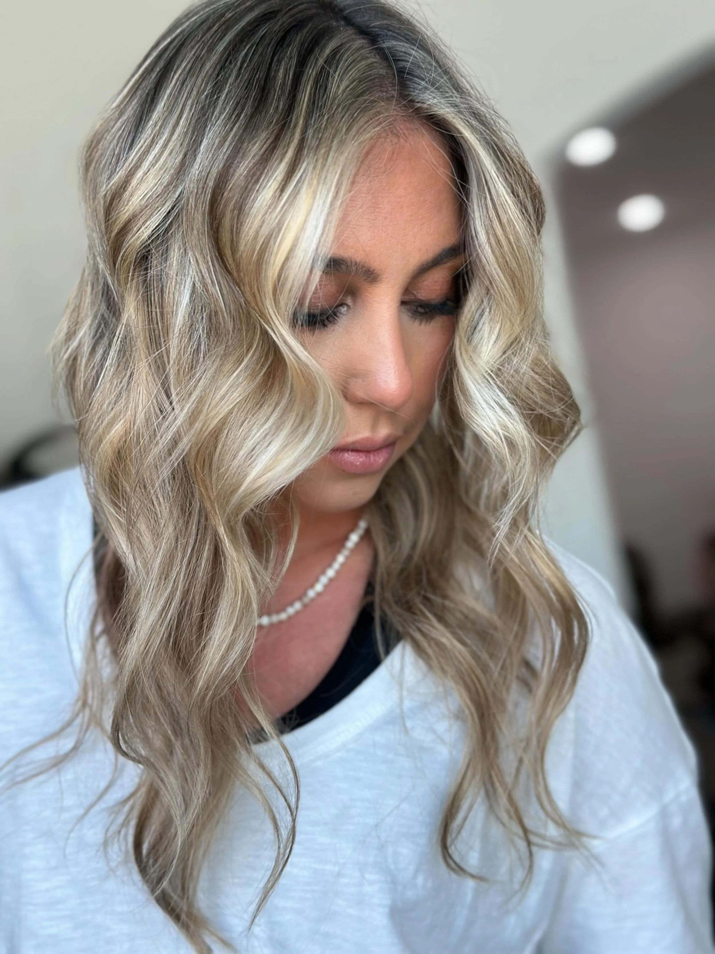 A woman with long, blonde, wavy hair looking down, wearing a white top and a pearl necklace, indoors with a blurred background and ceiling lights.