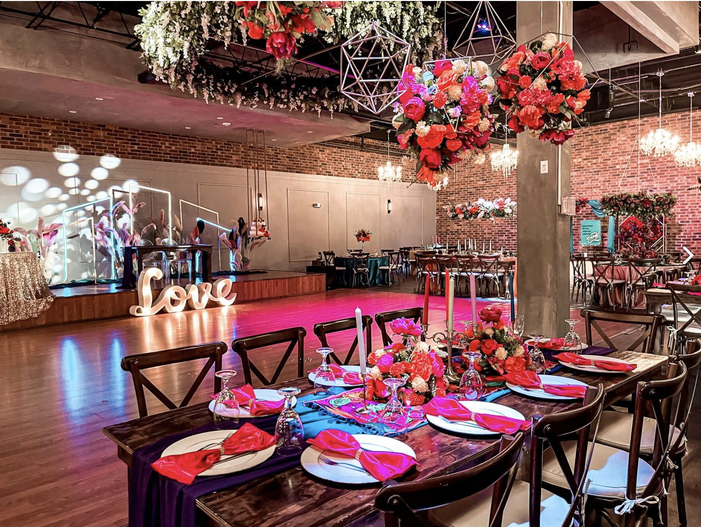 Reception setup featuring rustic wood tables, black chairs, red floral centerpieces, and hanging greenery.
