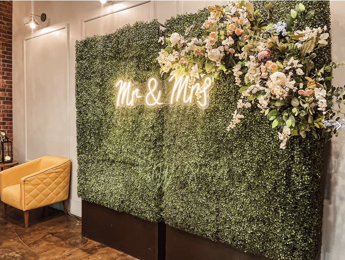 Greenery photo backdrop decorated with flowers, a neon “Mr & Mrs” sign, and a gold bench.